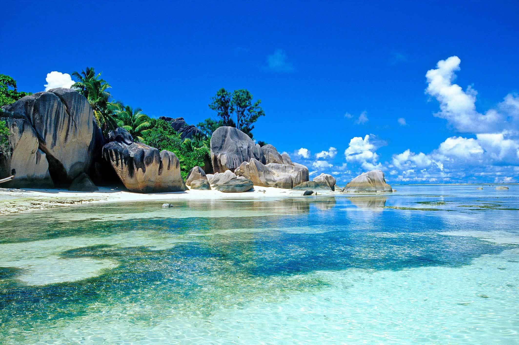 Beautiful Beach 桌布 壁紙 — 清澈碧綠的海水拍打著白色沙灘，沙灘四周環繞著巨大的花崗岩巨石和茂密的綠色棕櫚樹，上方是明亮的藍天和蓬鬆的白雲。