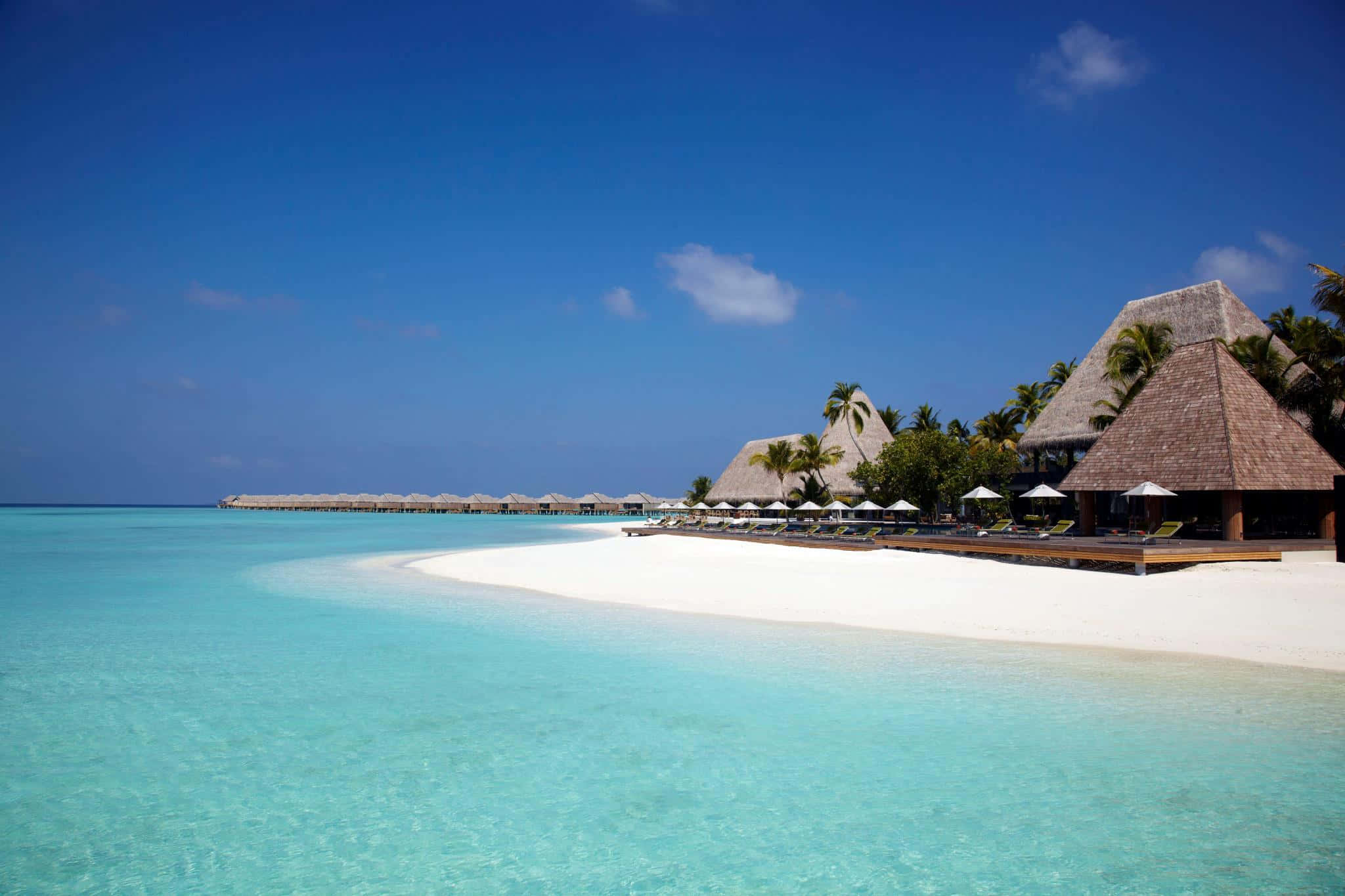Maldives Beach 桌布 壁紙 — 清澈碧綠的海水拍打著純淨的白色沙灘，沙灘上排列著茅草屋頂的水上平房，上方是湛藍的天空。