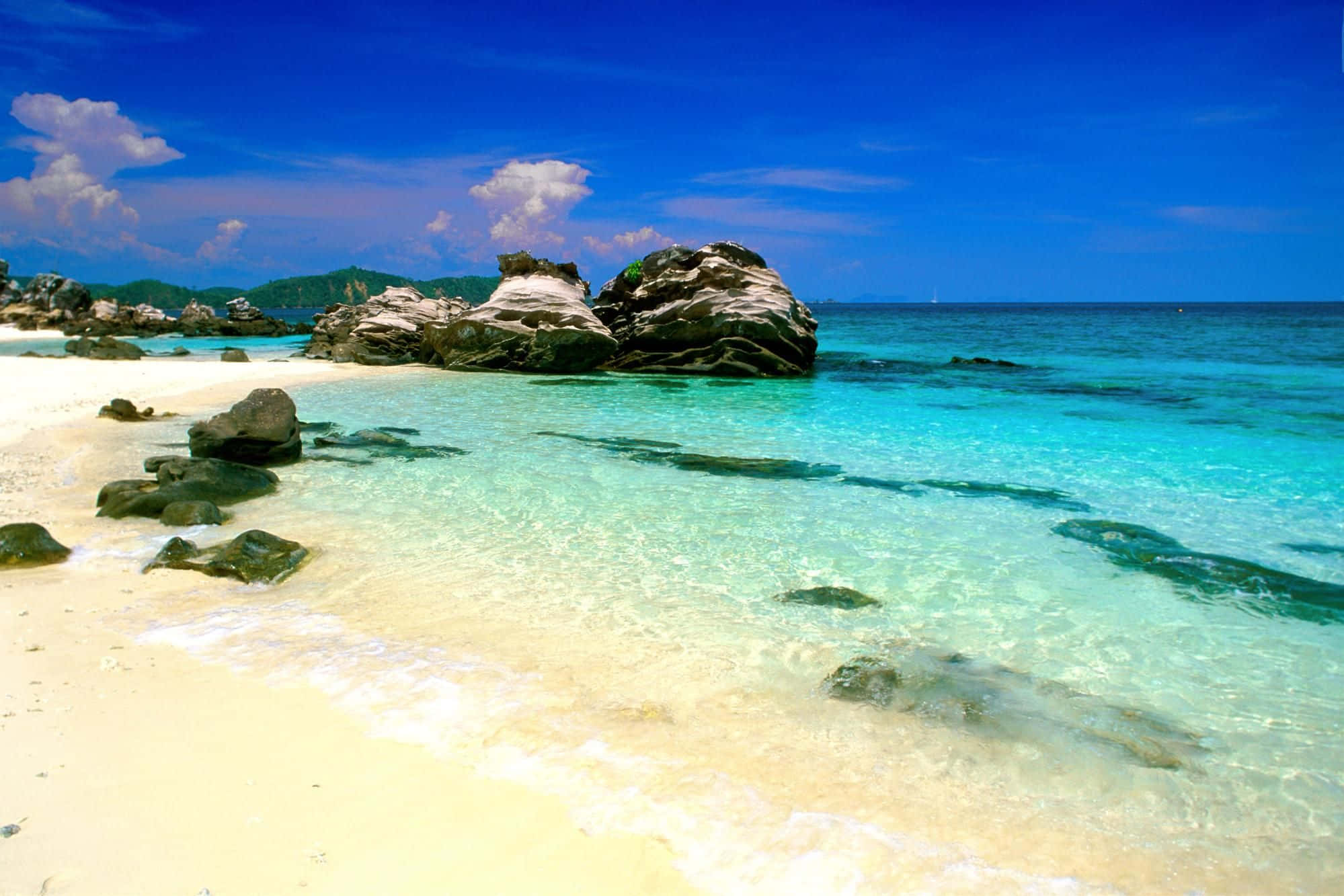 Thailand Beach 桌布 壁紙 — 清澈碧綠的海水拍打著純白沙灘，沙灘被戲劇性的深色岩石環繞，上方是鮮豔的藍天。
