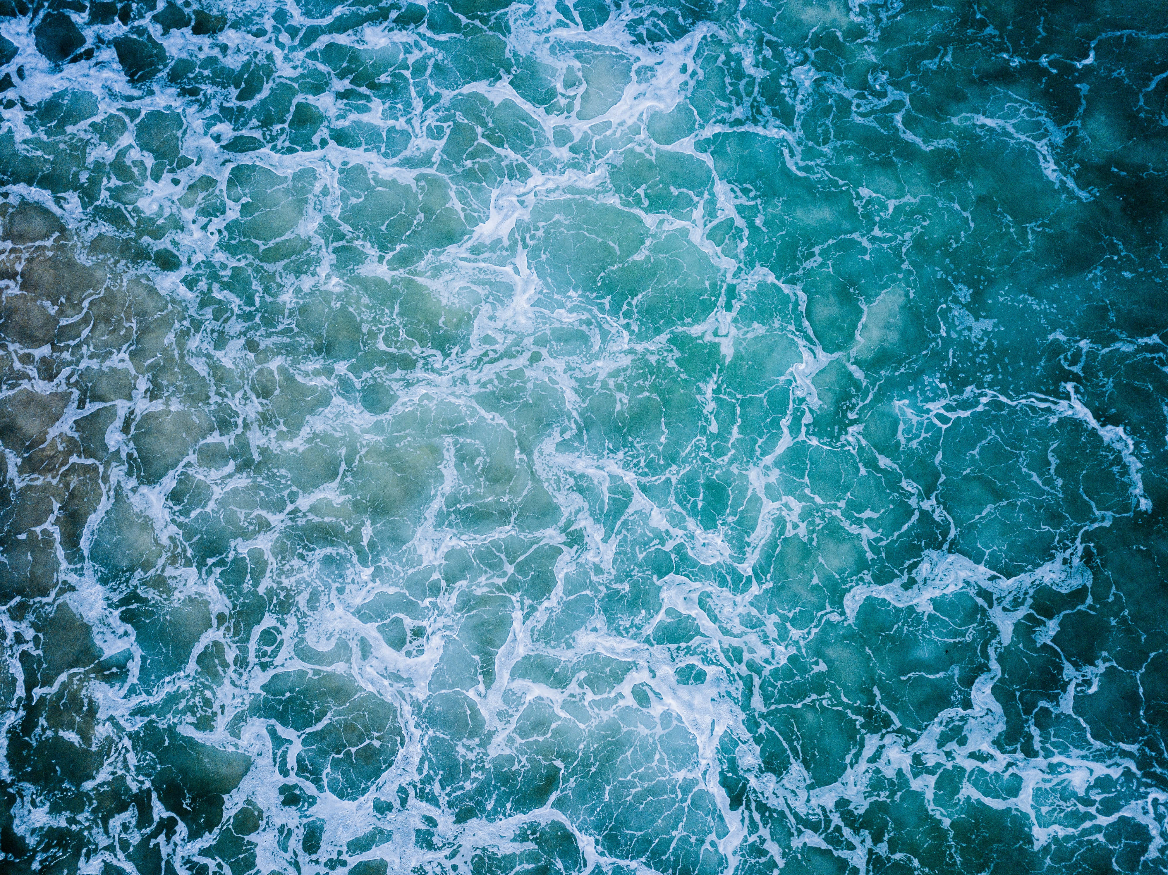 清澈藍色水紋 壁紙 — 從高空俯瞰，洶湧的藍綠色海浪翻騰，白色泡沫狀浪花形成複雜的漩渦圖案。