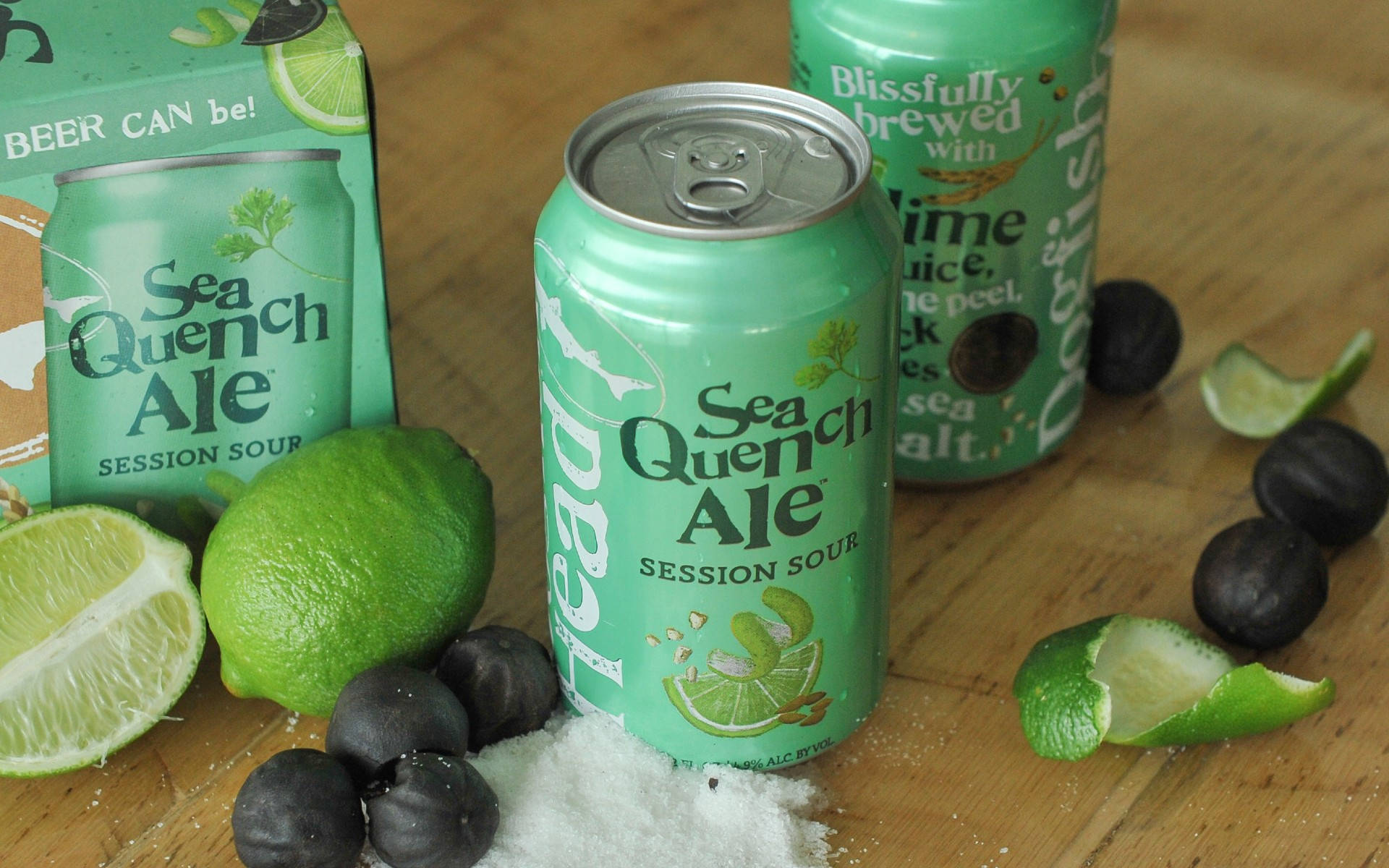 清爽檸檬汽水罐裝飲品 壁紙 — 一罐充滿活力的綠色 Sea Quench Ale 啤酒，周圍環繞著青檸、鹽和黑橄欖，放在木質表面上。