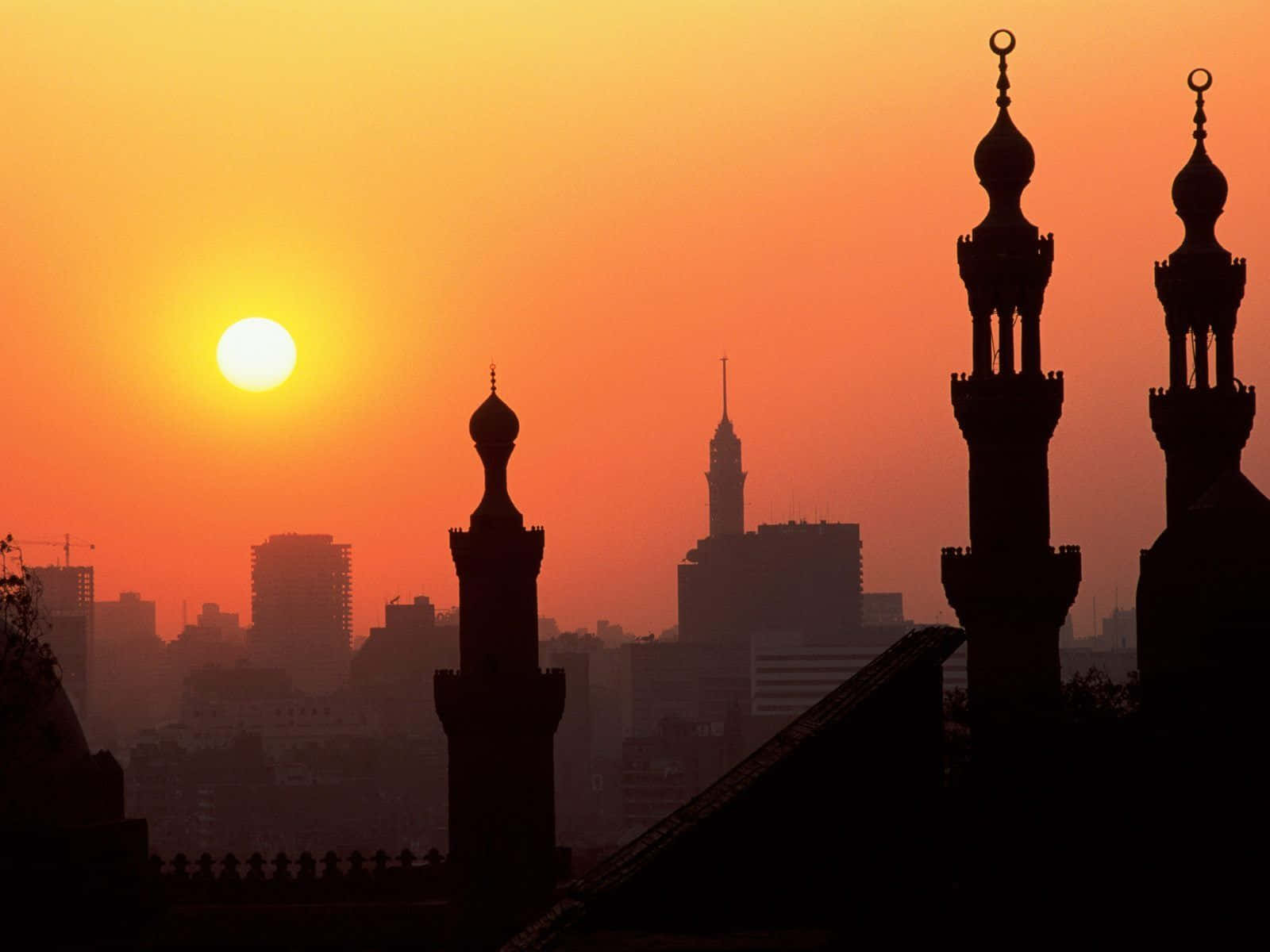 Egypt 桌布 壁紙 — 清真寺宣禮塔的剪影在火紅的橙色日落中升起，映襯著朦朧的城市天際線和一輪明亮的太陽。