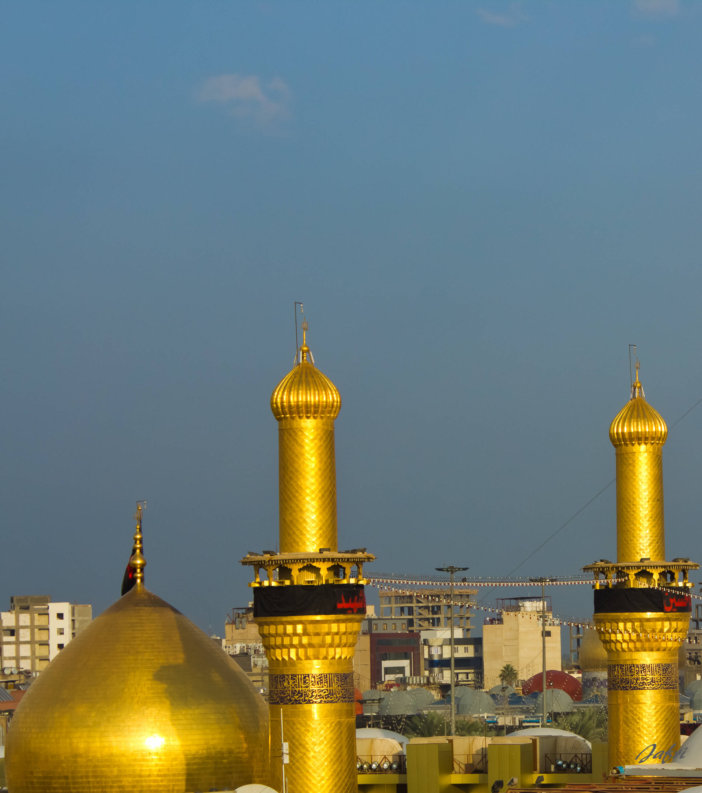 Karbala 桌布 壁紙 — 清真寺的金色圓頂和宣禮塔在晴朗的藍天下閃閃發光，背景是城市建築。