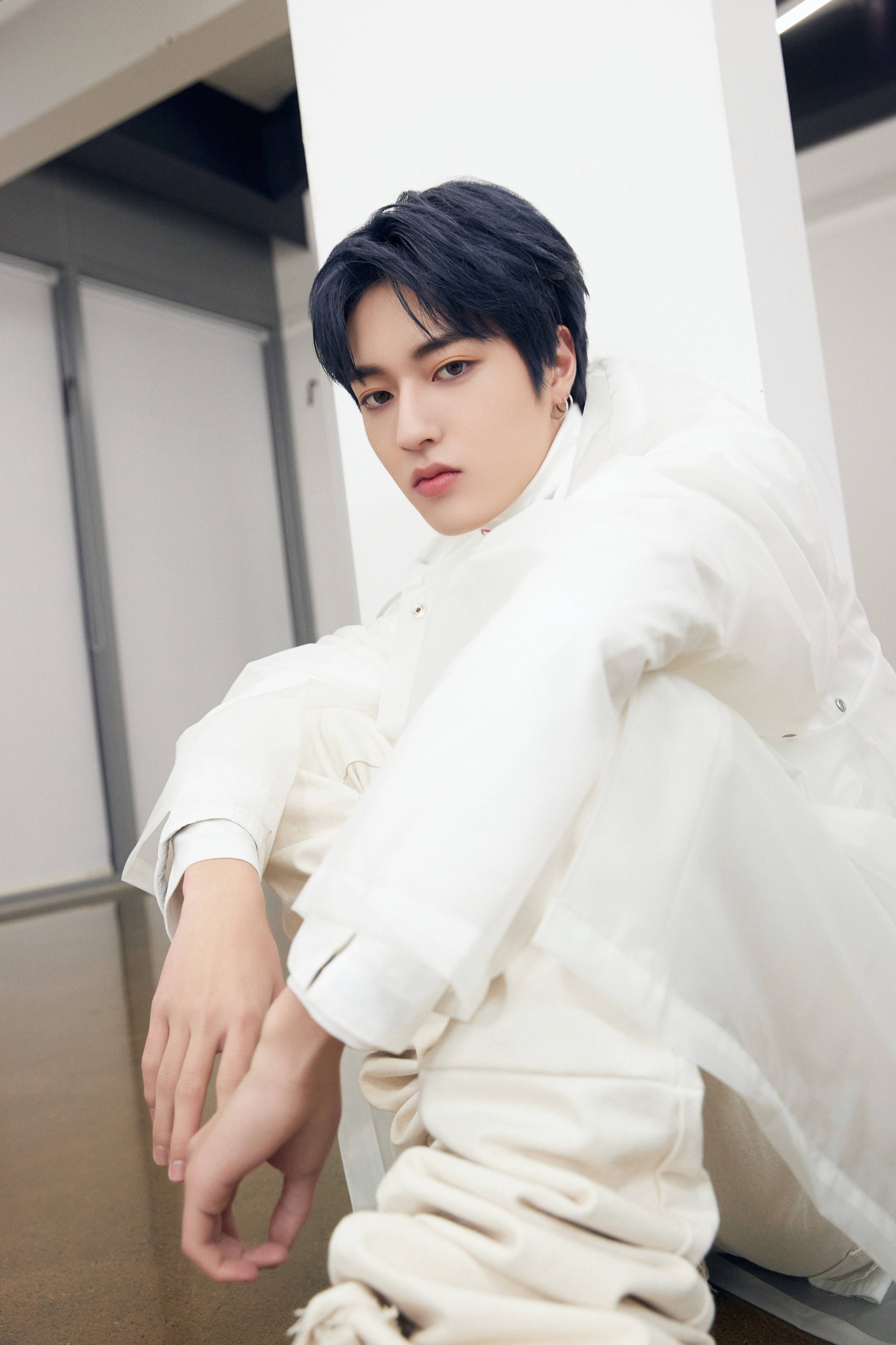 渡邊一馬 K-POP個性風 壁紙 — 一位深色頭髮的年輕男子坐在現代室內空間，靠著白色柱子，身穿寬鬆白色服裝。