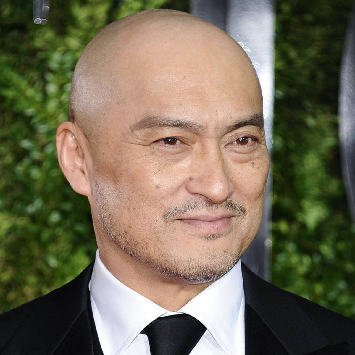 穿黑色西裝打領帶的禿頭男人在活動中對著鏡頭微笑。