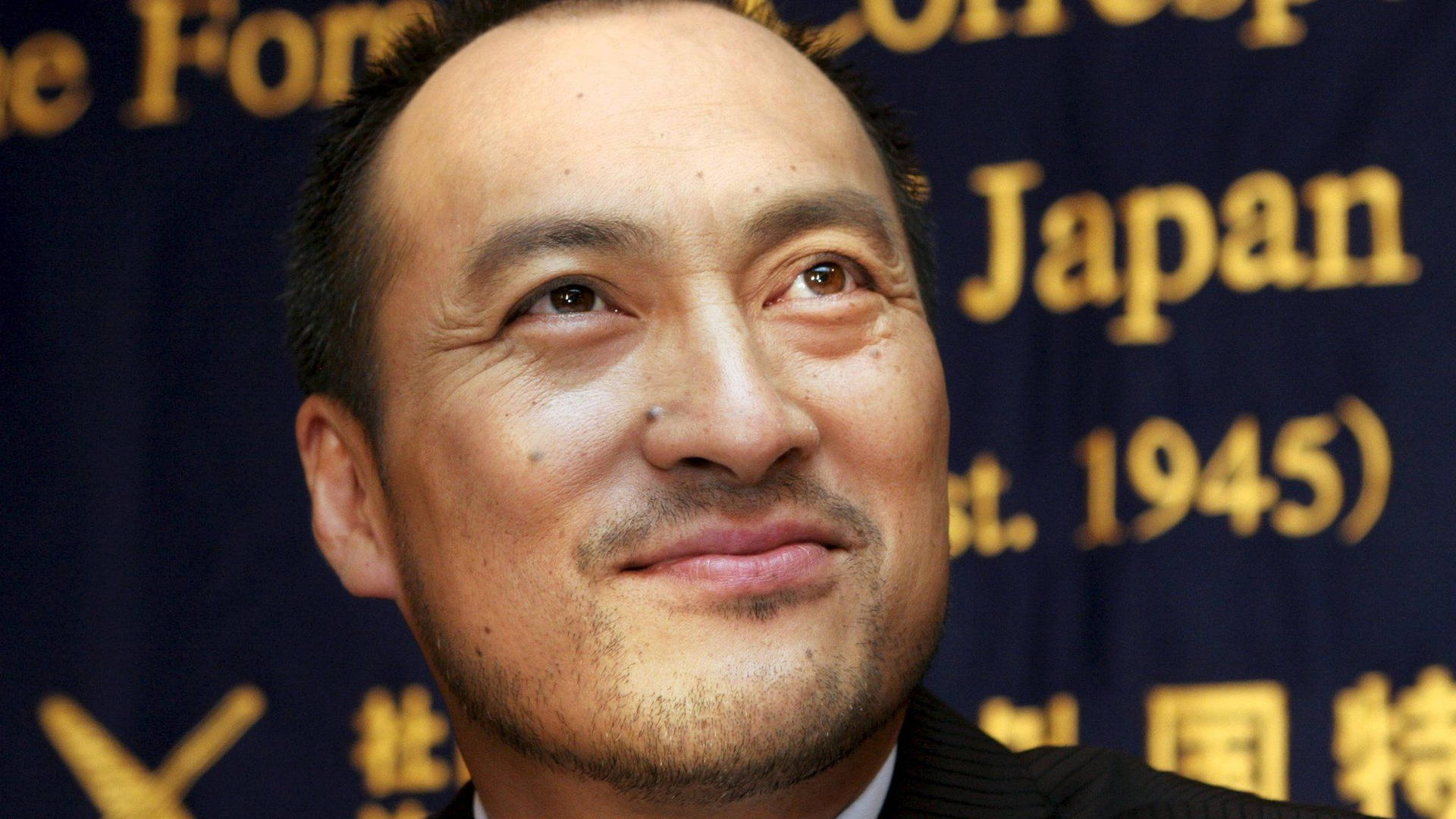 日本活動中身穿西裝微笑的男子，身後有金色字母。