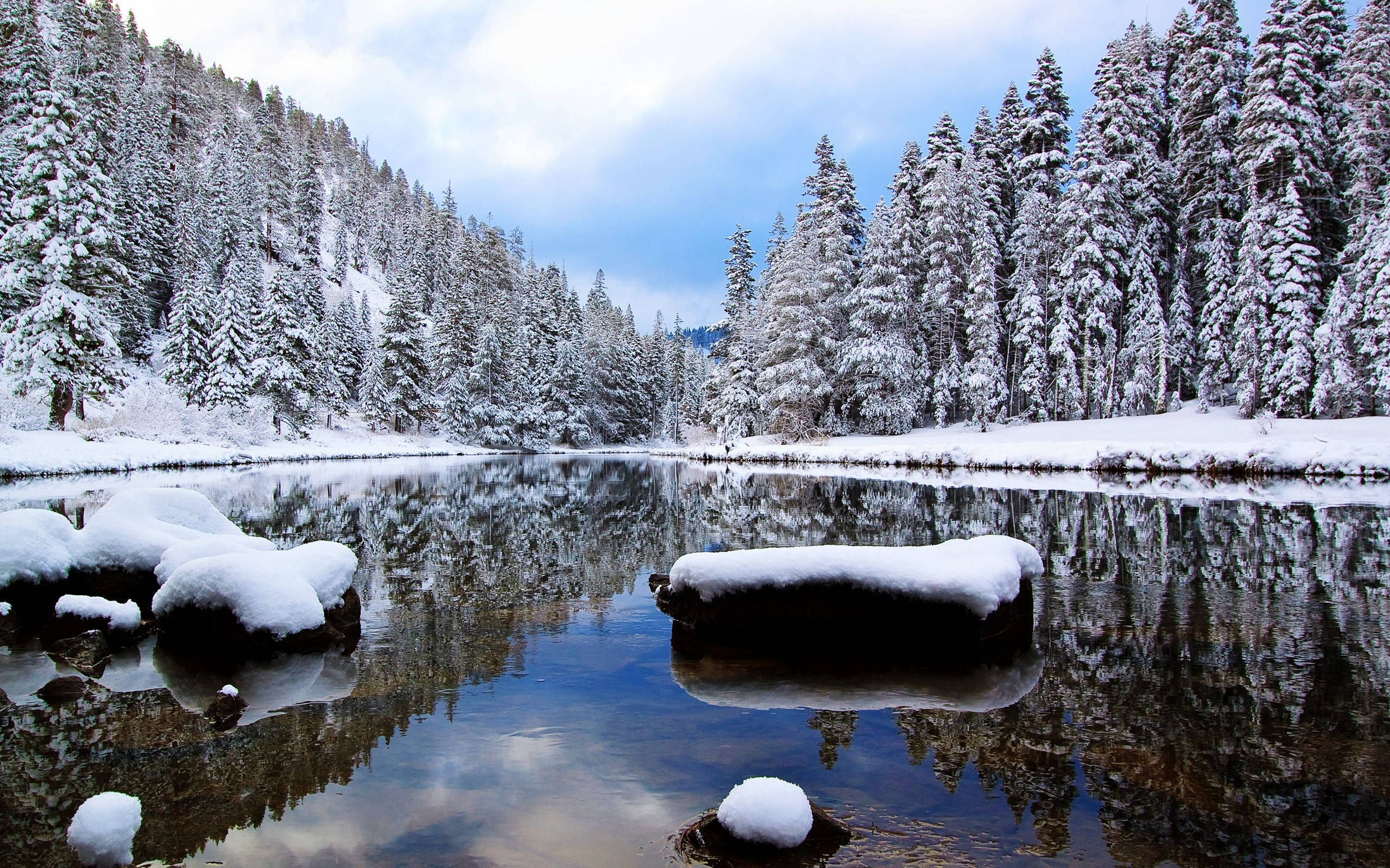 湖畔冬日風景 壁紙 — 白雪覆蓋著常綠樹和岩石，環繞著一個平靜的湖泊，倒映著多雲的冬日天空。