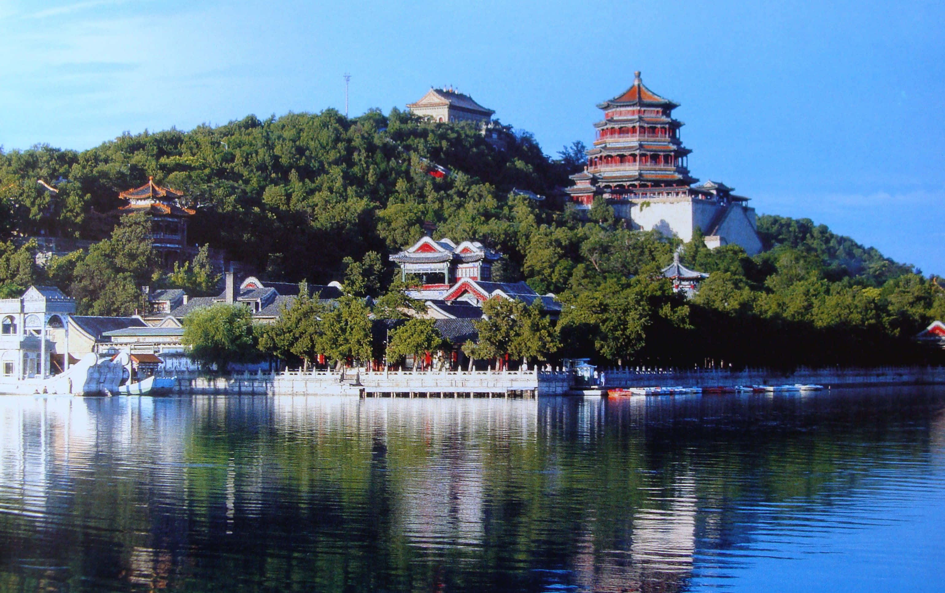 湖畔夏宮 壁紙 — 傳統中式寶塔和亭台樓閣坐落在鬱鬱蔥蔥的青山上，在晴朗的天空下完美地倒映在平靜的藍色水面上。