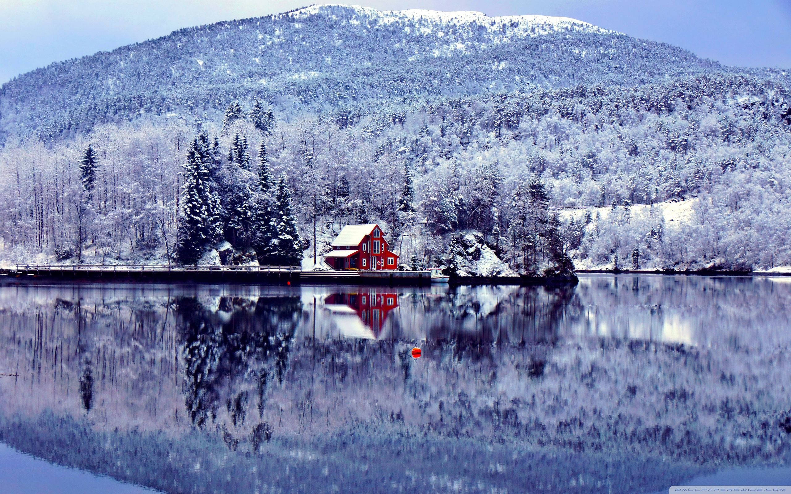 湖畔小屋冬日風景 壁紙 — 一間鮮紅色的木屋坐落在白雪覆蓋的湖岸邊，在平靜的水面上完美倒映，周圍是白雪覆蓋的常綠樹木和山脈。