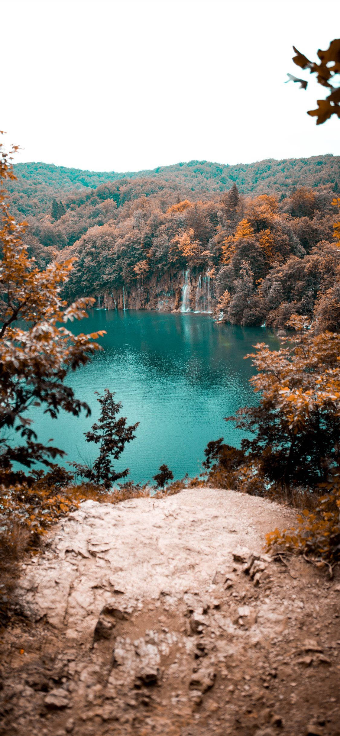 湖畔瀑布風景 壁紙 — 寧靜的綠松石色湖泊隱藏在秋日樹林中，瀑布傾瀉入平靜的水面，四周環繞著金色的樹葉。