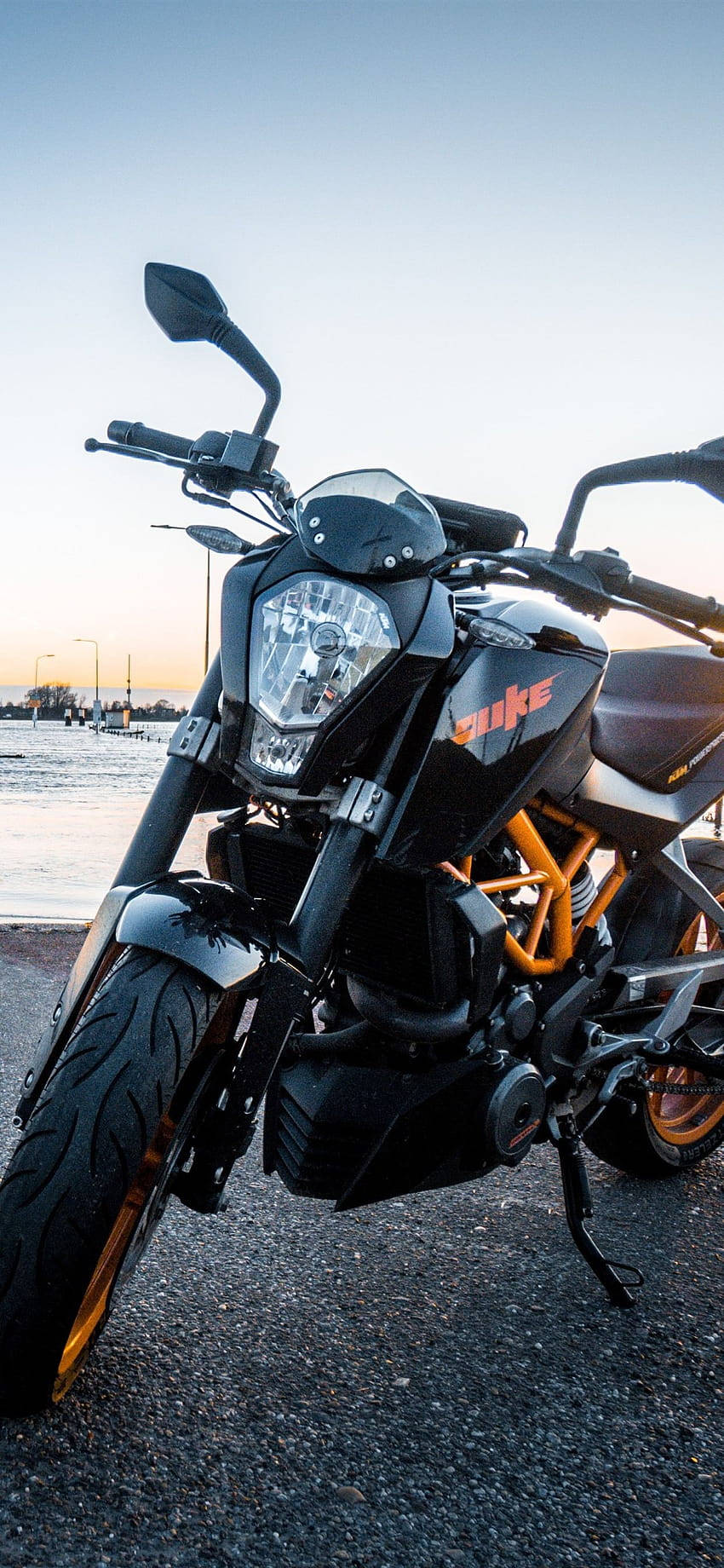 黑色KTM Duke電單車帶橙色細節，停泊在日落時分近水邊。