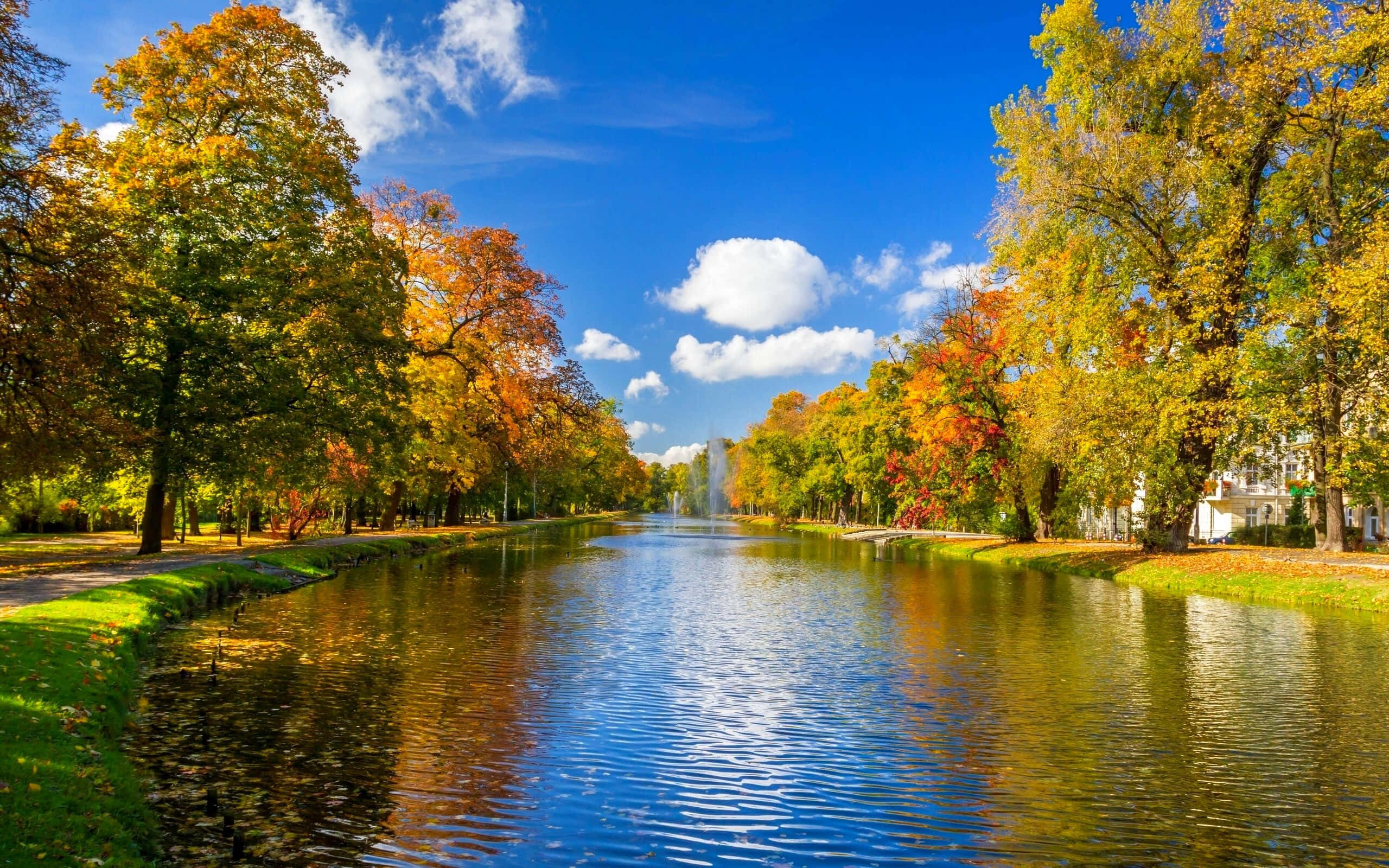 湖畔秋日公園與樹木 壁紙 — 一條寧靜的秋天運河，兩旁是鮮豔的橙色和黃色樹木，頭頂是明亮的藍天和蓬鬆的白雲。