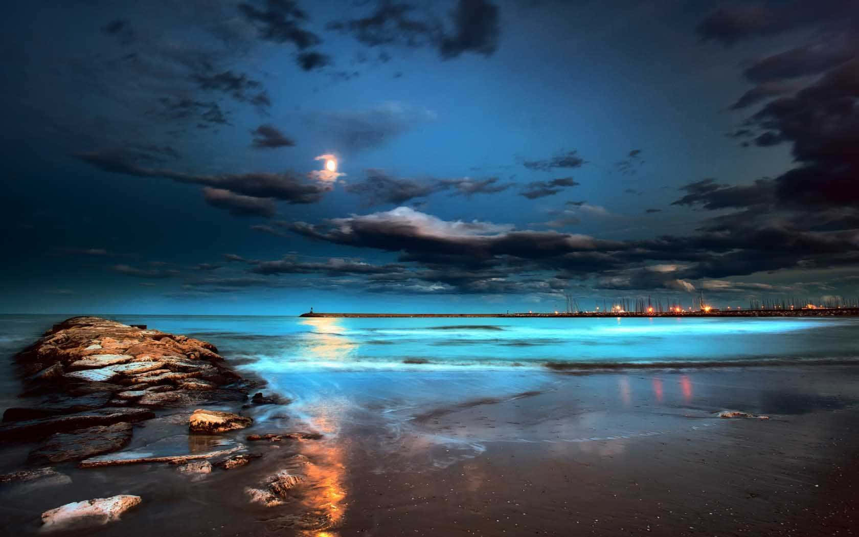 多雲夜空下，月光照耀的海灘，有青綠色的海浪和黑色岩石。