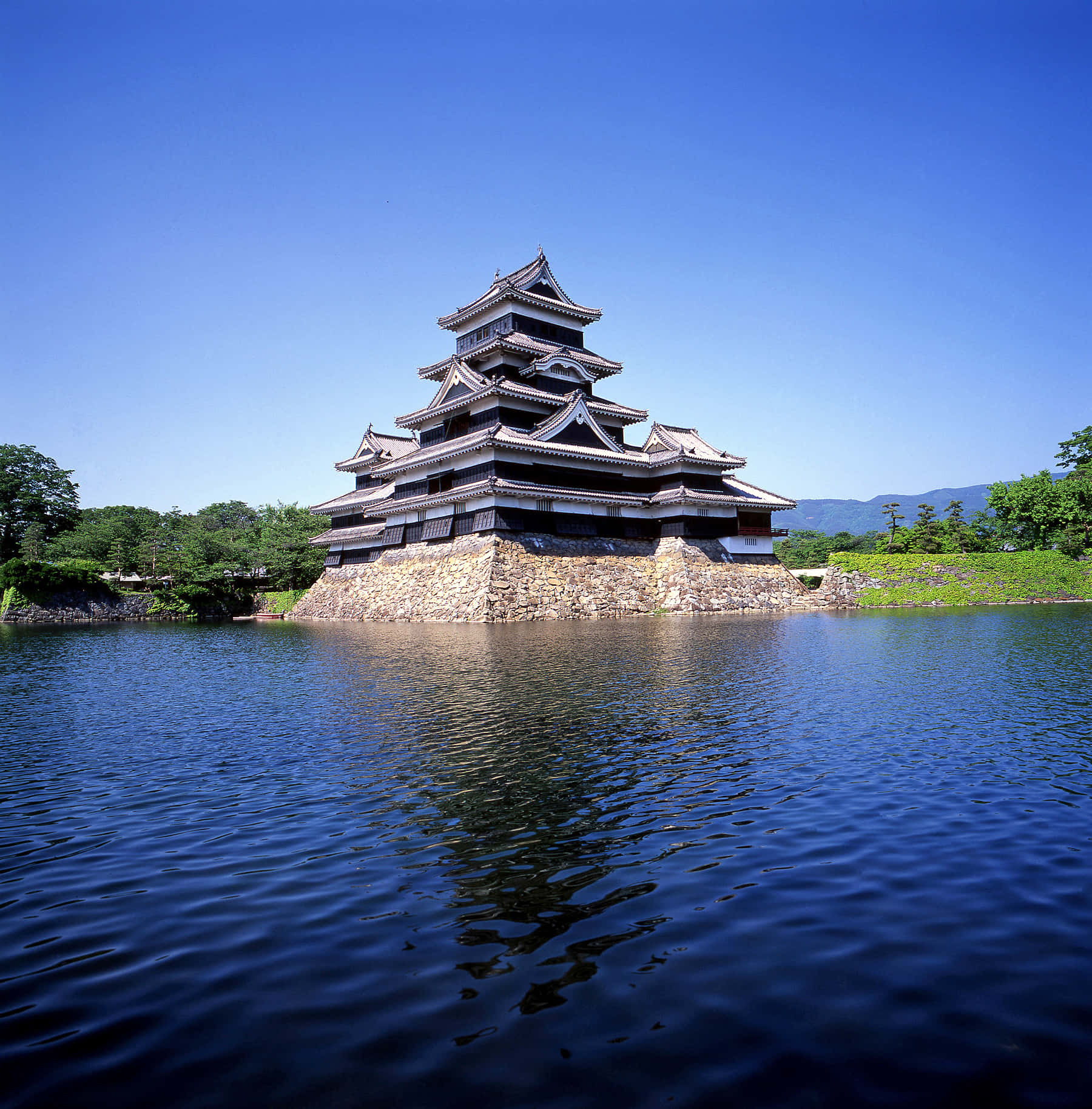 湖畔美麗寺廟風景 壁紙 — 一座雄偉的傳統日本城堡矗立在石基上，周圍環繞著平靜的深色水域，上方是清澈的藍天。