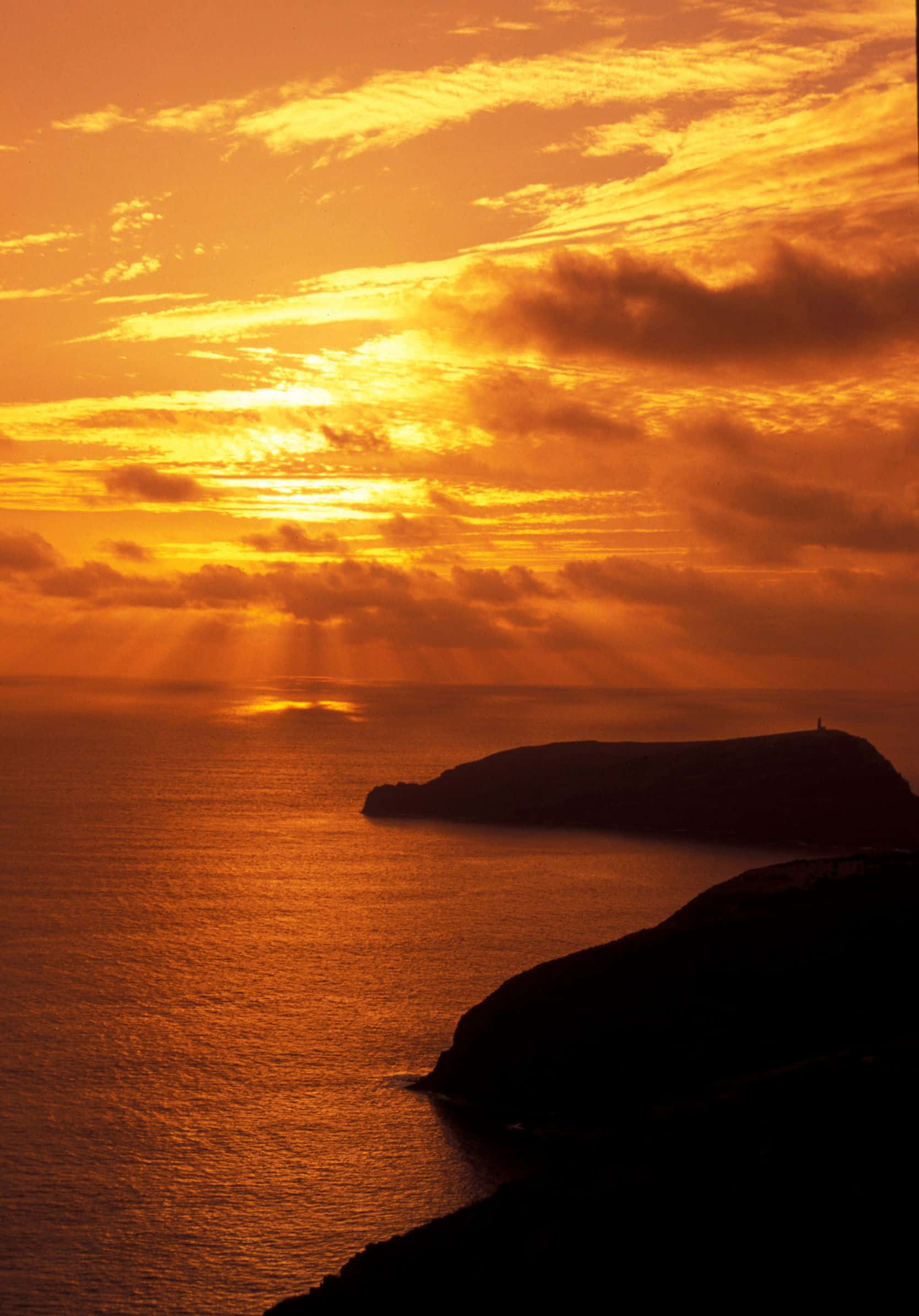 湖畔靜賞夕陽 壁紙 — 金色陽光穿透火橙色雲層，照耀在平靜的海洋上，懸崖在日落時形成剪影。