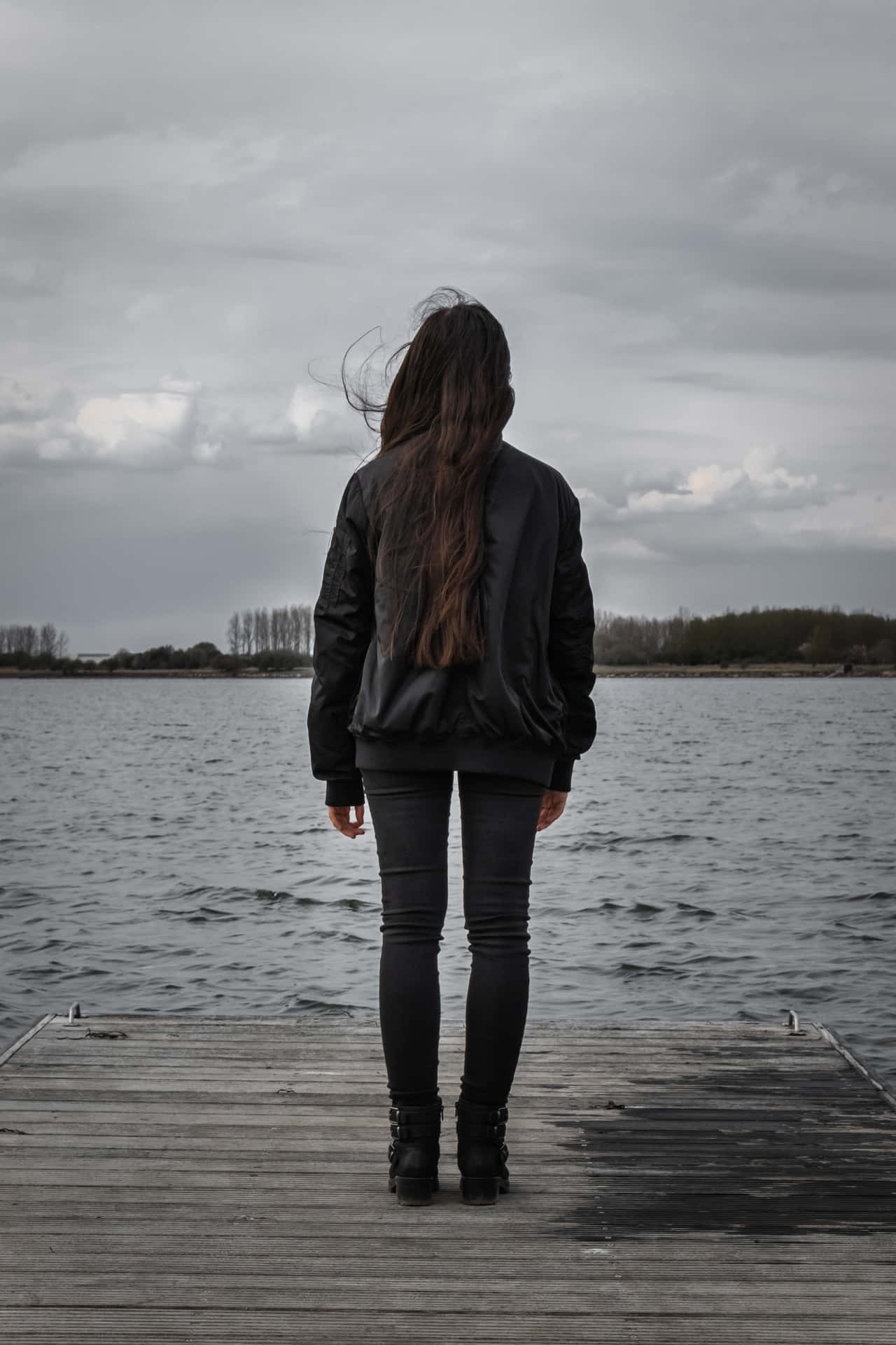 穿著黑色外套的女人獨自站在木碼頭上，俯瞰陰天下平靜的湖泊。