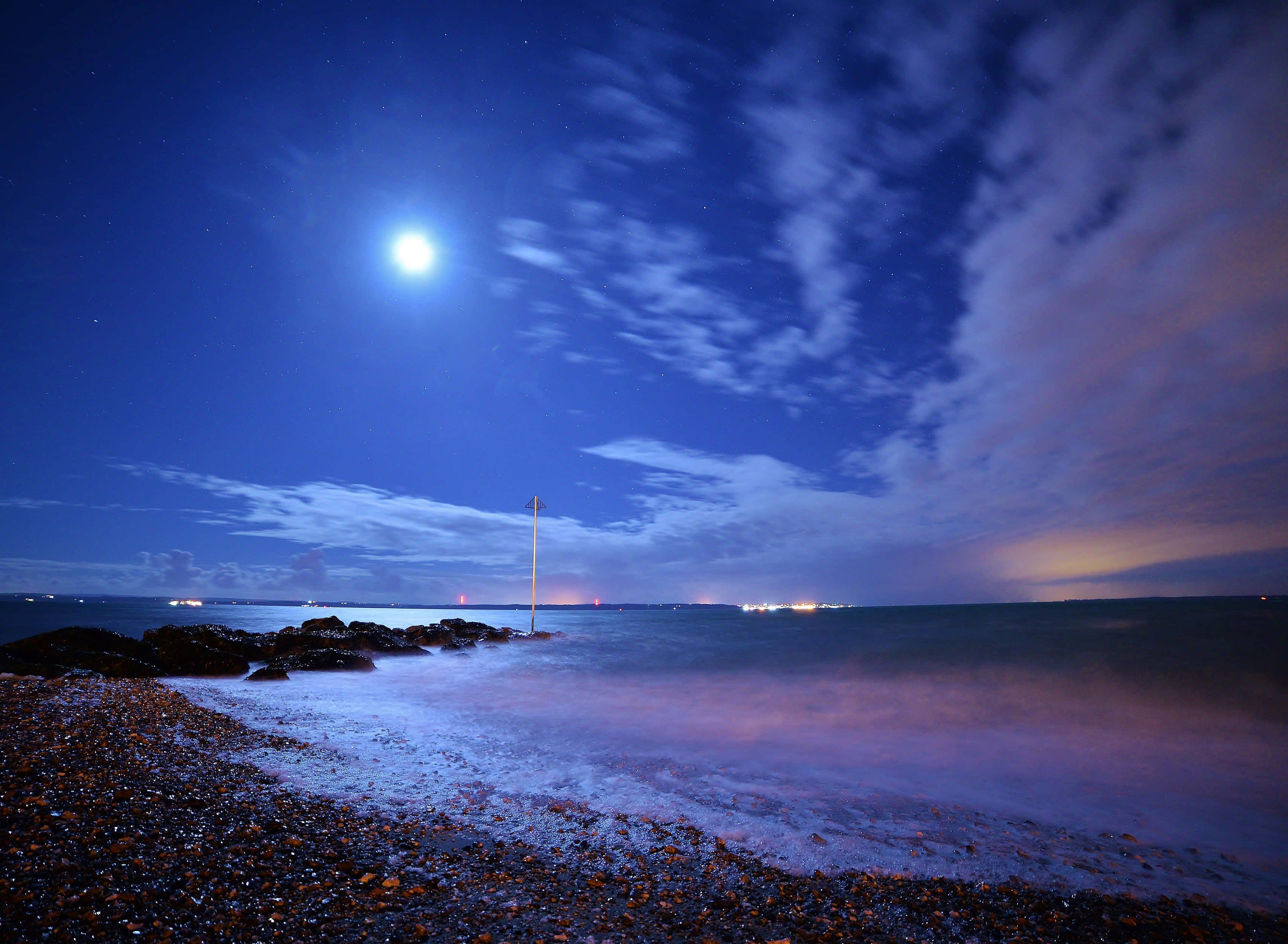 湛藍夜空海灘圖 壁紙 — 一輪明亮的滿月在卵石海灘上投下寧靜的光芒，海浪在繁星點點的暮色天空下拍打。