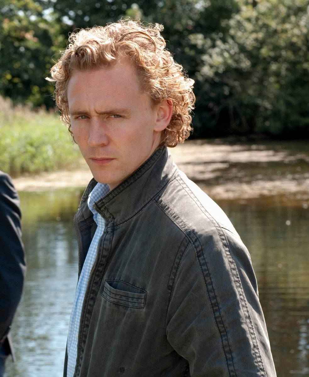 湯姆·希德勒斯頓出演《Wallander》 壁紙 — 一個捲曲金髮、身穿深色外套的男人站在平靜的河邊，在明亮的日光下，背景有樹木。
