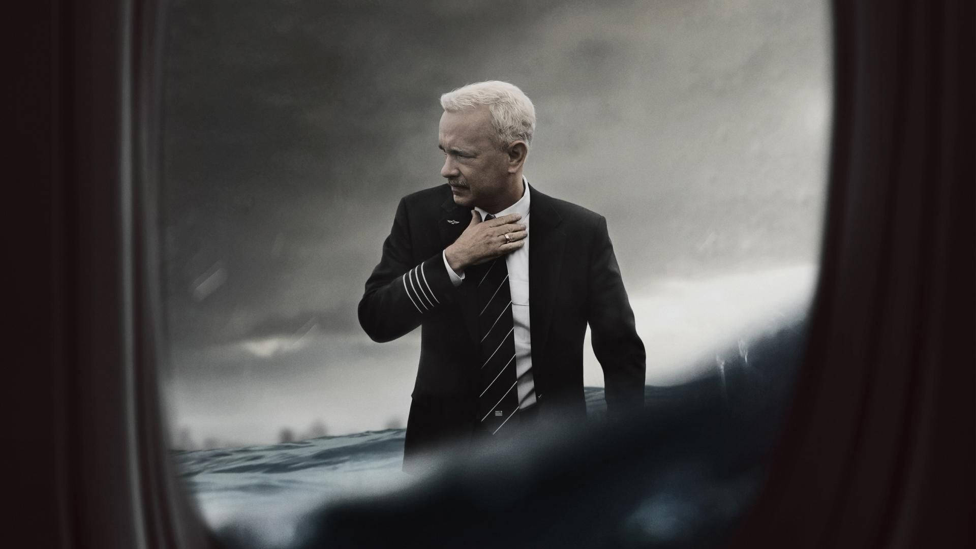 湯姆·漢克斯《薩利機長》 壁紙 — 一名身穿深色制服嘅機師喺暴風雨嘅海邊莊嚴咁企喺度，透過窗戶同模糊嘅前景窗簾望出去。