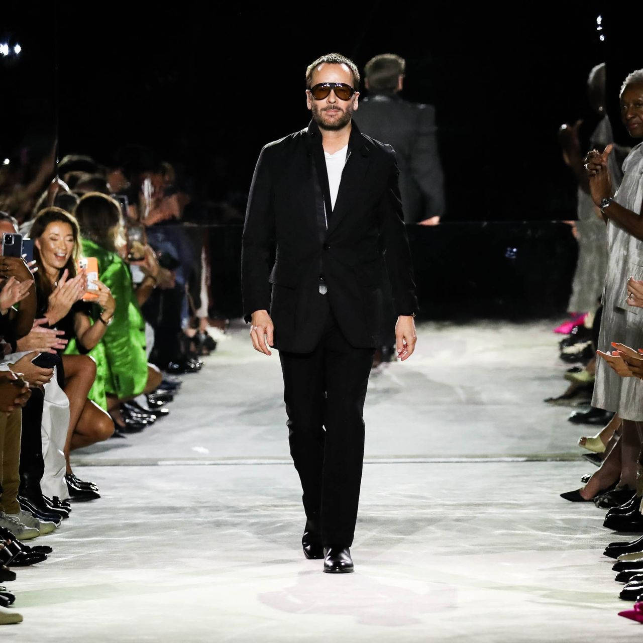 身穿黑色西裝和太陽眼鏡的男子在鼓掌的觀眾面前走 runway。