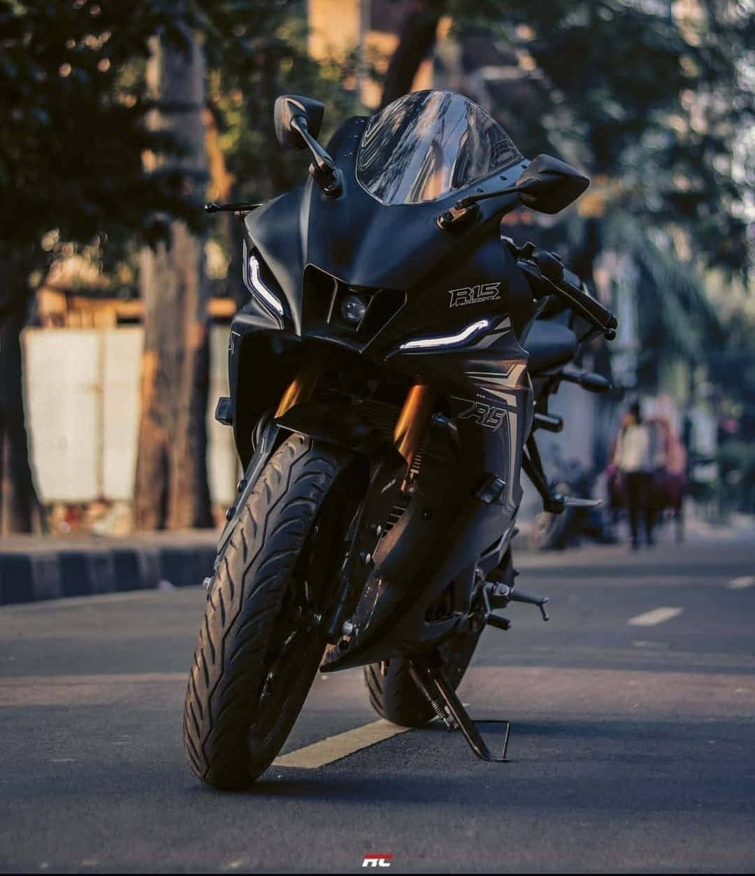 準備在賽道上與 Yamaha R15 競速 壁紙 — 一架時尚黑色電單車停泊在樹木林立的城市街道上，沐浴在金色的日落光線中。