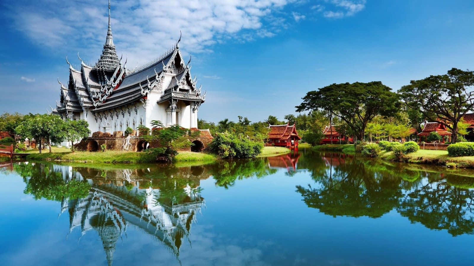 白色泰式寺廟倒映在平靜的池塘中，周圍有綠樹和藍天