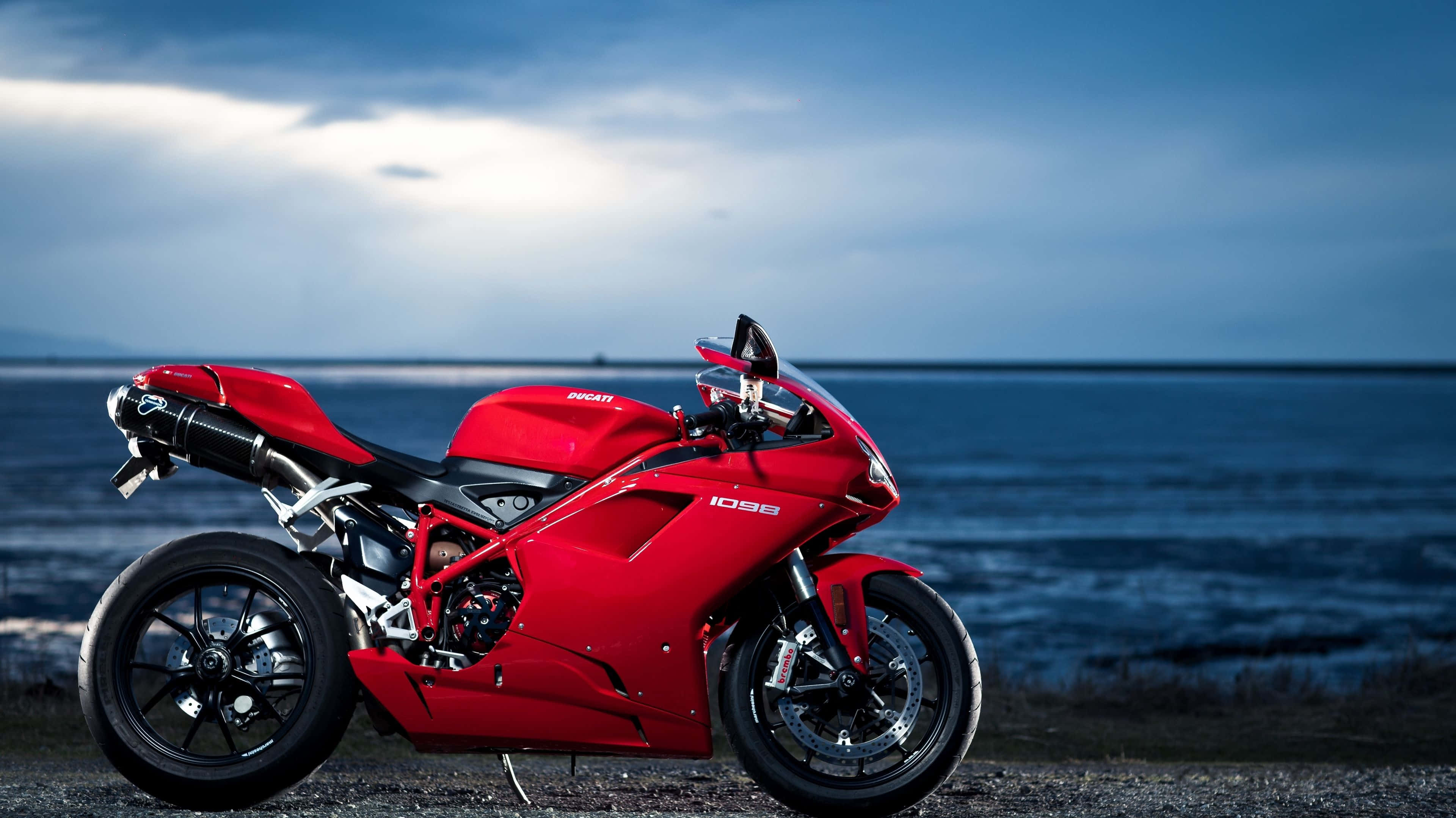 準備好與4K自行車一同出發 壁紙 — 一架搶眼的紅色Ducati 1098電單車停在礫石岸邊，背景是廣闊的藍色海洋和陰鬱的黃昏天空。