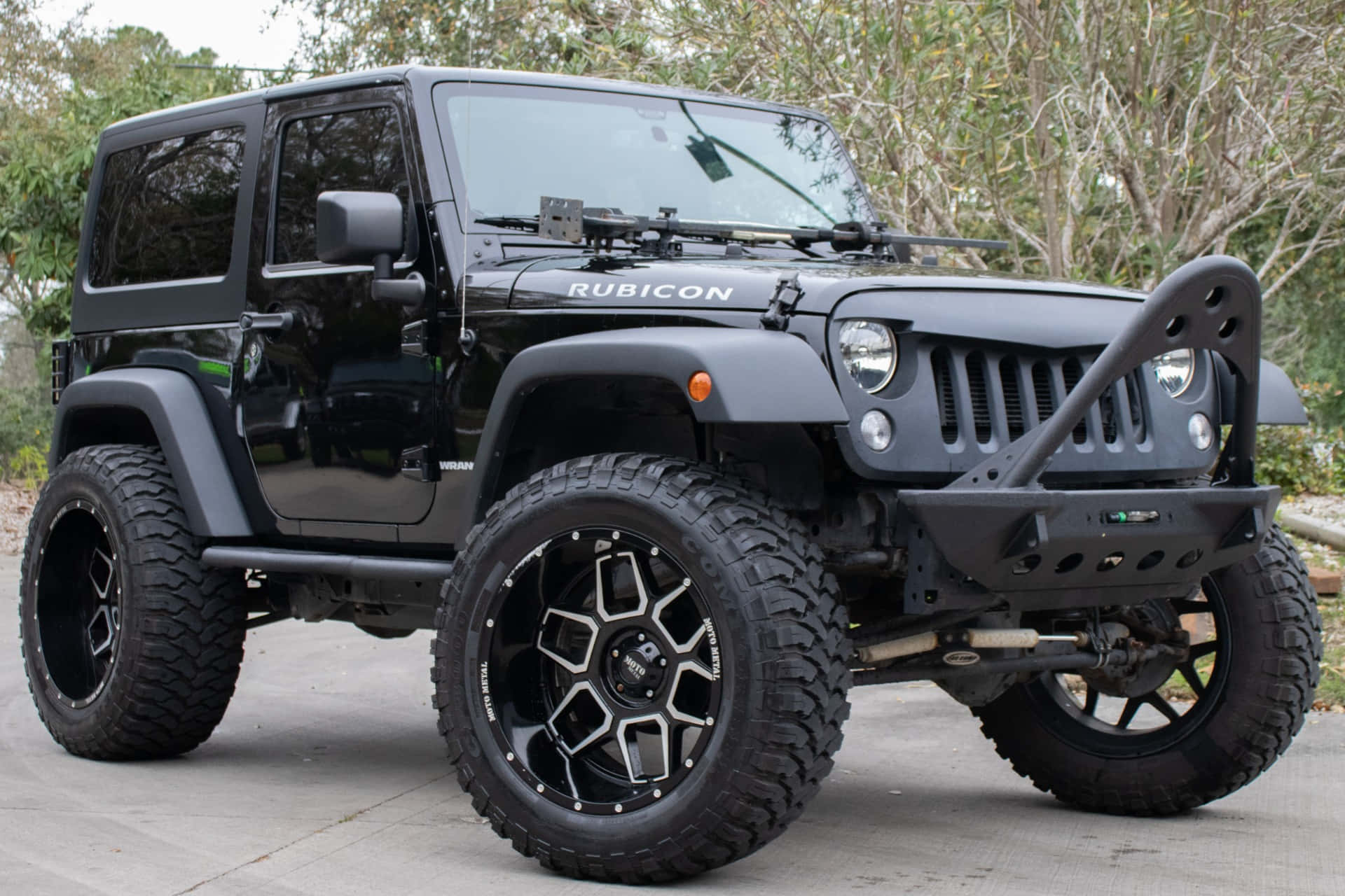 準備好與經典Jeep Wrangler一同迎接每一天的冒險 壁紙 — 一輛啞光黑色Jeep Wrangler Rubicon配備超大輪胎和定制輪圈，停放在乾樹環繞的混凝土表面上。
