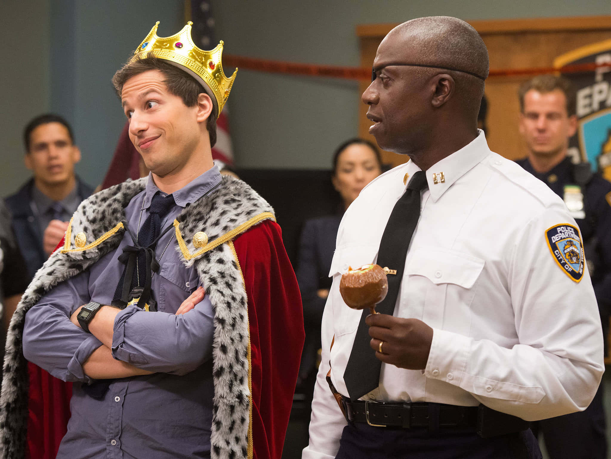 準備好迎接歡笑吧！《Brooklyn Nine-Nine》來了！ 壁紙 — 一名戴著金色王冠和紅色斗篷的男子在法庭上，旁邊是一名拿著冬甩的警察，他露出得意的笑容。