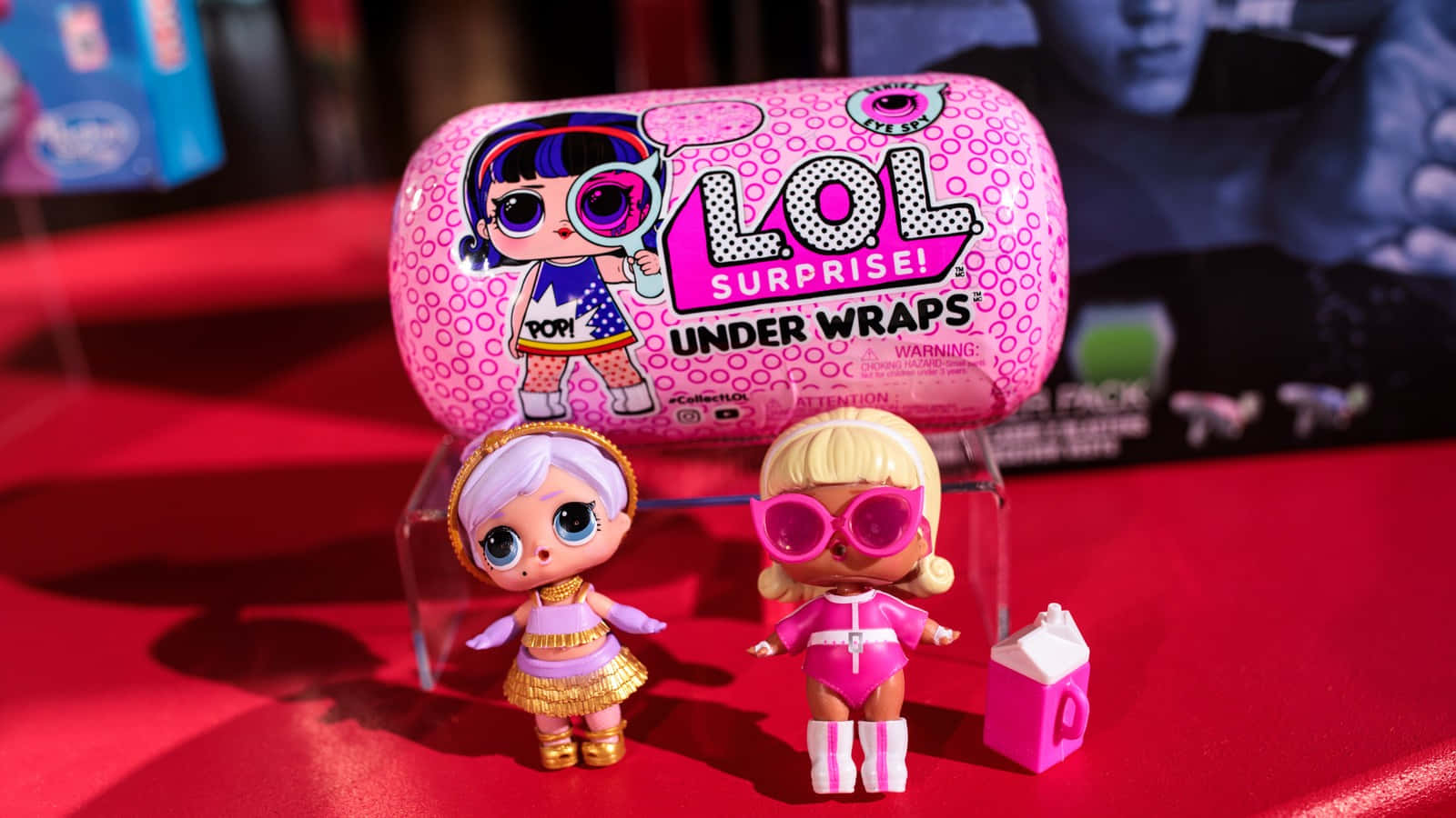準備探索LOL娃娃的奇妙世界！ 壁紙 — 兩個身穿粉紅和金色服裝的LOL Surprise公仔站在紅色表面上，前面是它們的粉紅色圓柱形玩具盒。