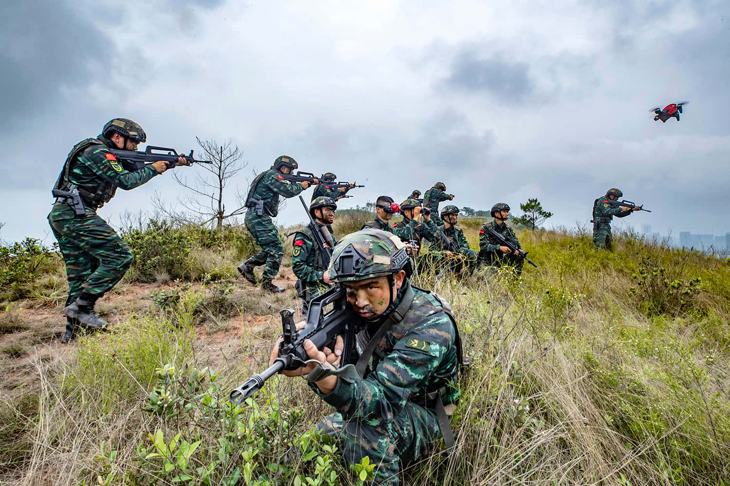 準備迎戰 壁紙 — 身穿迷彩裝備的士兵在陰天天空下穿越草坡，一架無人機在頭頂飛過。