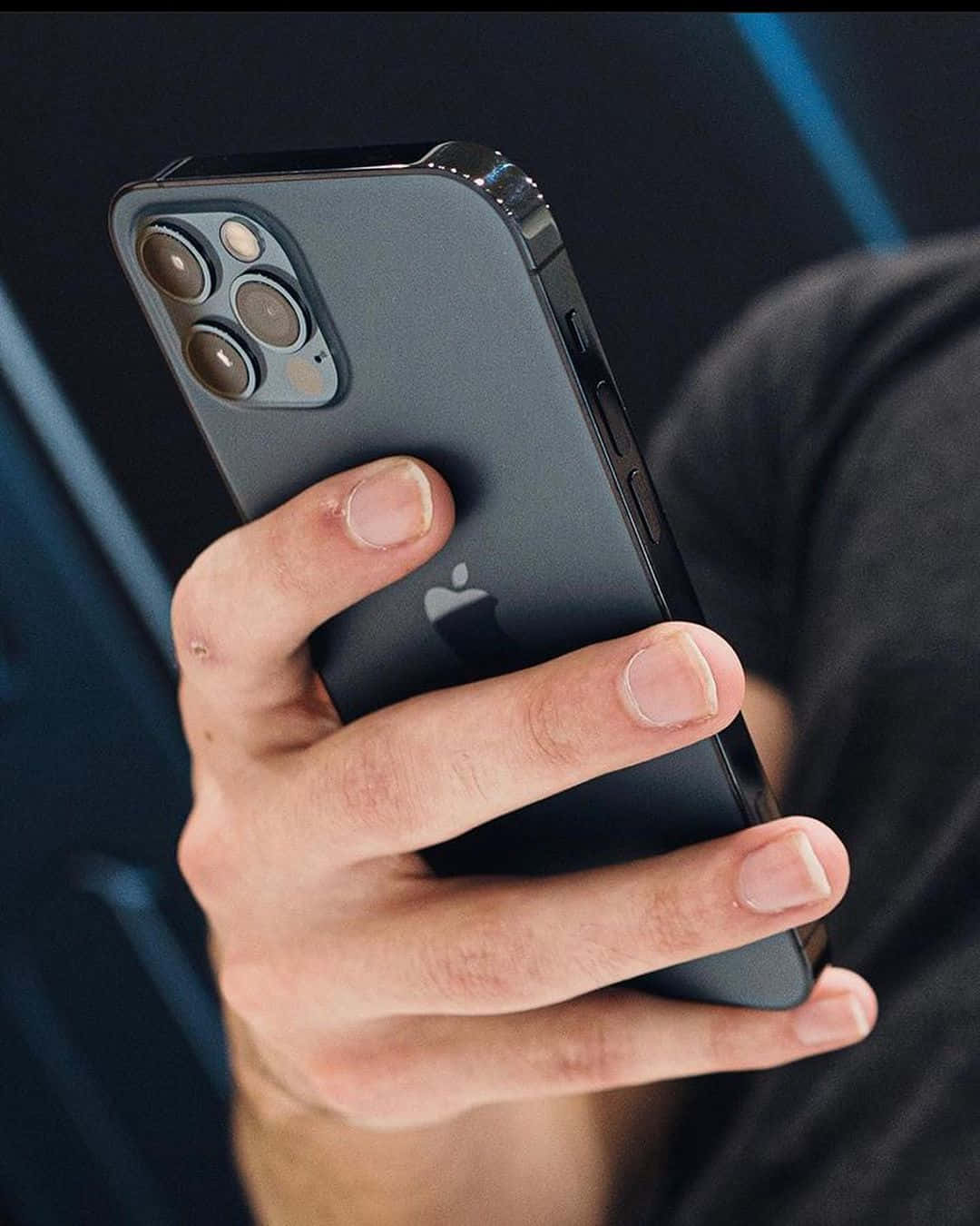 準備迎接全新 iPhone 12 壁紙 — 一隻手緊握深灰色iPhone 12 Pro的特寫，其三鏡頭系統在模糊的藍色背景下清晰可見。