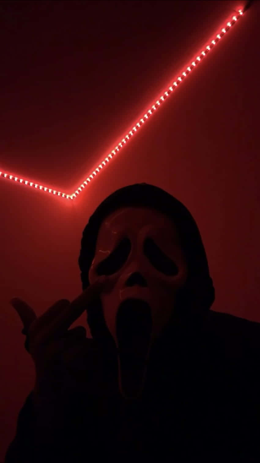 戴著Ghostface面具嘅人喺紅色燈光嘅房間入面指向相機。