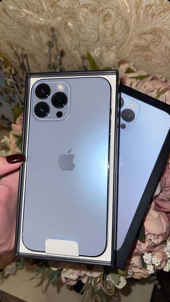 準備迎接最新 iPhone！ 壁紙 — 一隻手拿著一個新的淺藍色 iPhone 13 Pro 放在盒子裡，周圍環繞著柔和的粉紅色花朵和華麗的壁紙。