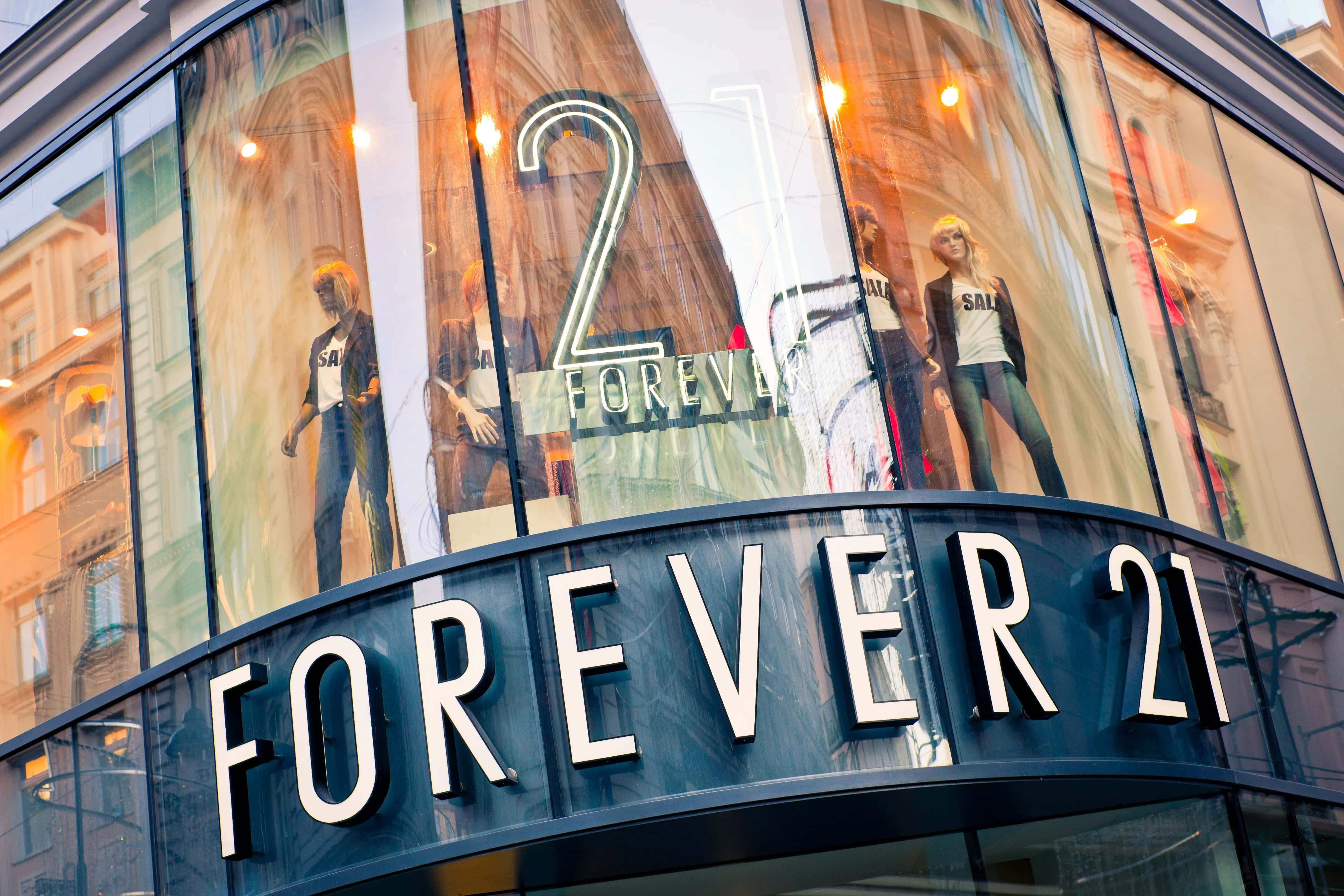 準備迎接最酷的季節，盡情展現自我風格 — Forever 21 壁紙 — Forever 21 的弧形玻璃店面展示穿著時尚服裝的人體模型，在溫暖的室內燈光下。