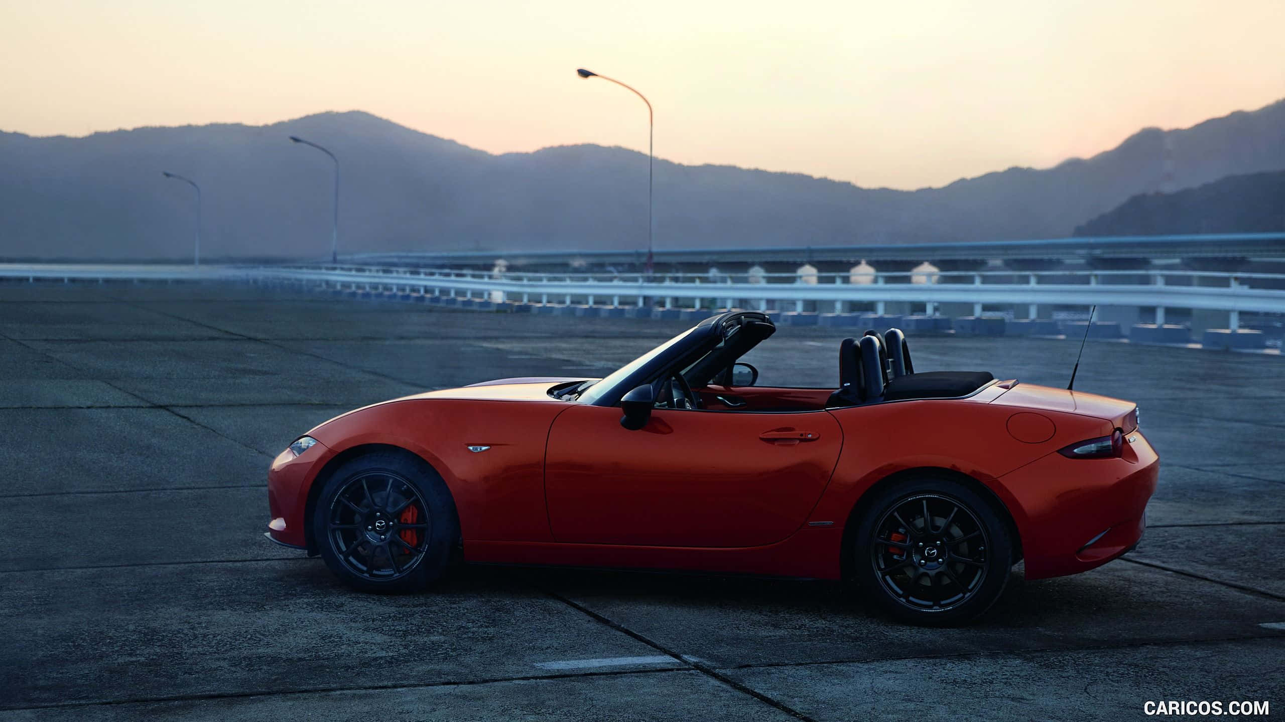 準備迎接難忘駕駛體驗，馬自達MX-5 Miata 壁紙 — 一架充滿活力的橙色開篷跑車在日落時停泊在高速公路橋上，背景有山巒。