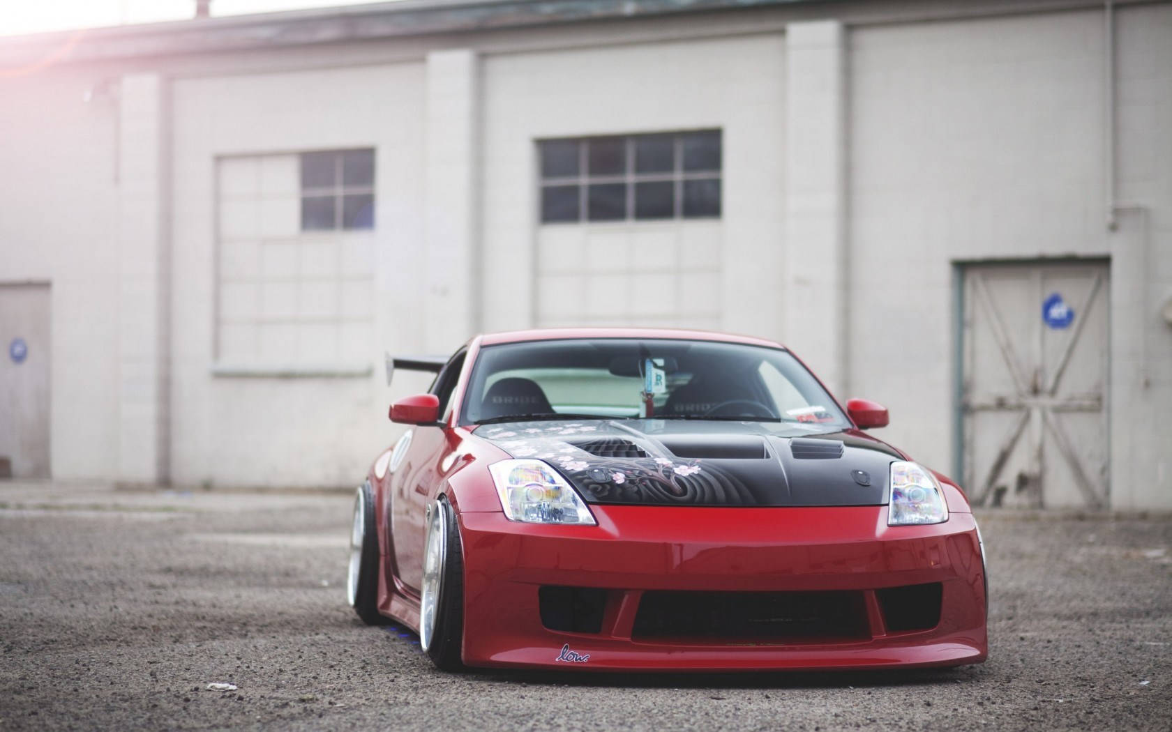 「準備駕駛 Nissan 350Z」 壁紙 — 一架光面紅色改裝跑車，黑色引擎蓋和定制輪圈，停泊在風化的工業建築前。