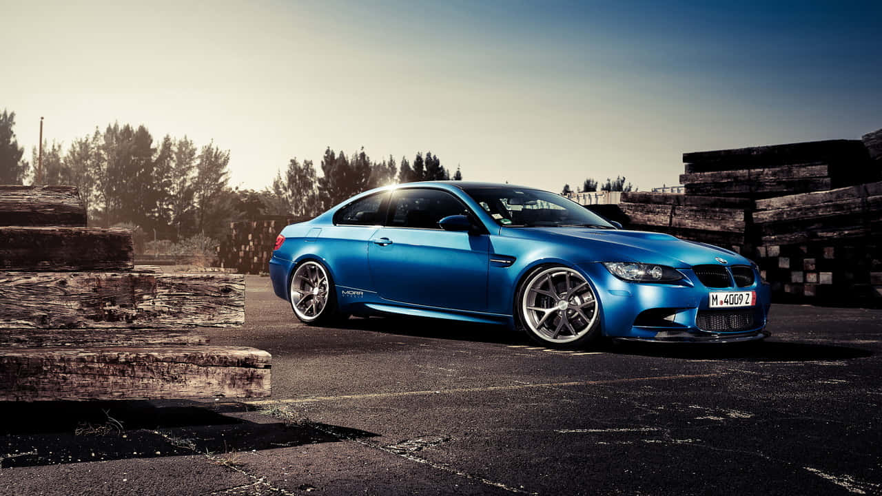 準備駕駛BMW，享受奢華旅程 壁紙 — 一輛充滿活力的藍色BMW M3雙門跑車在黃金時段的光線下閃閃發光，停放在堆疊的木樑旁邊。