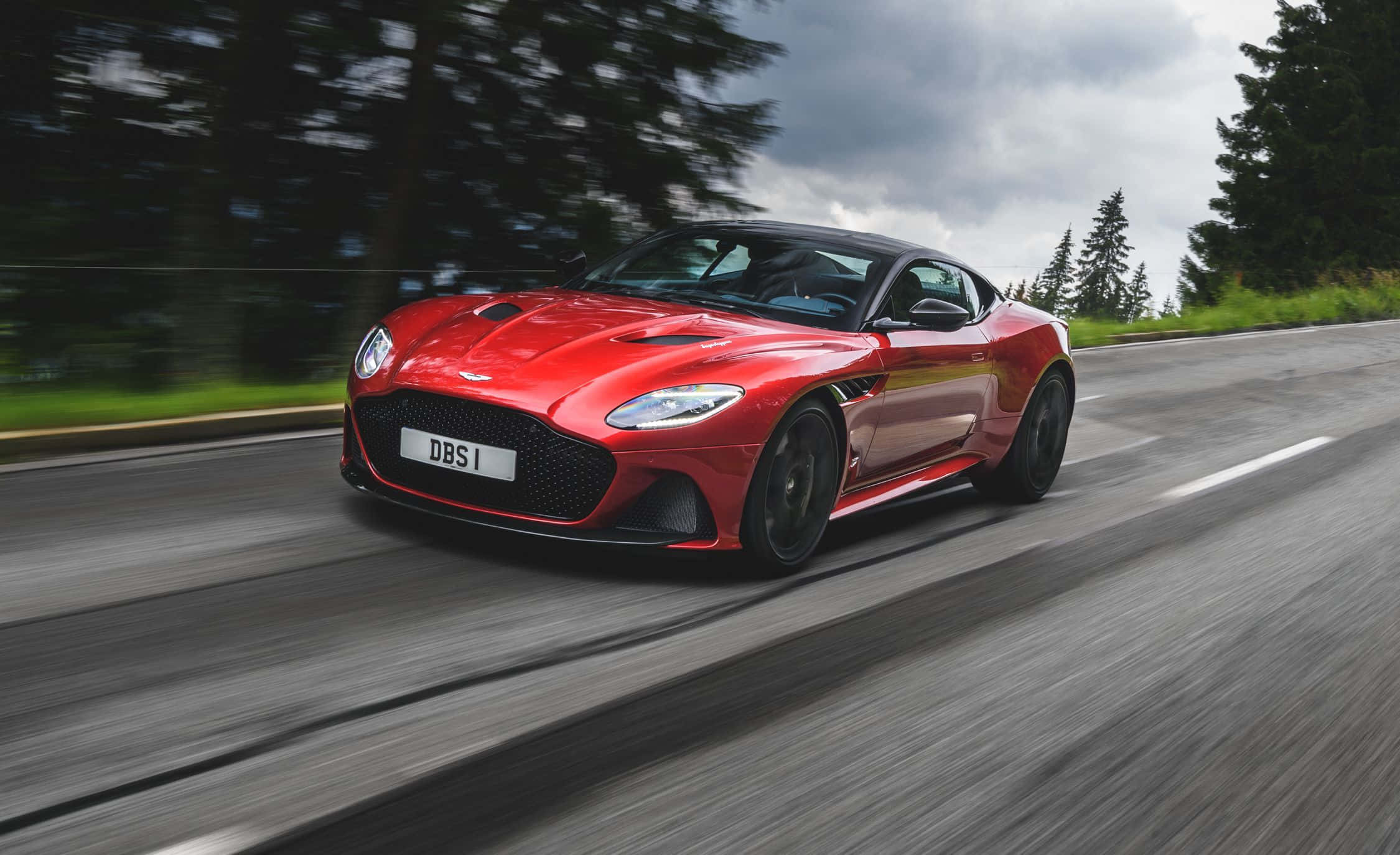 準備體驗阿斯頓·馬丁的高性能駕駛 壁紙 — 一輛鮮紅色的Aston Martin DBS1超級跑車沿著蜿蜒的林間道路疾馳，動態模糊強調了其速度。