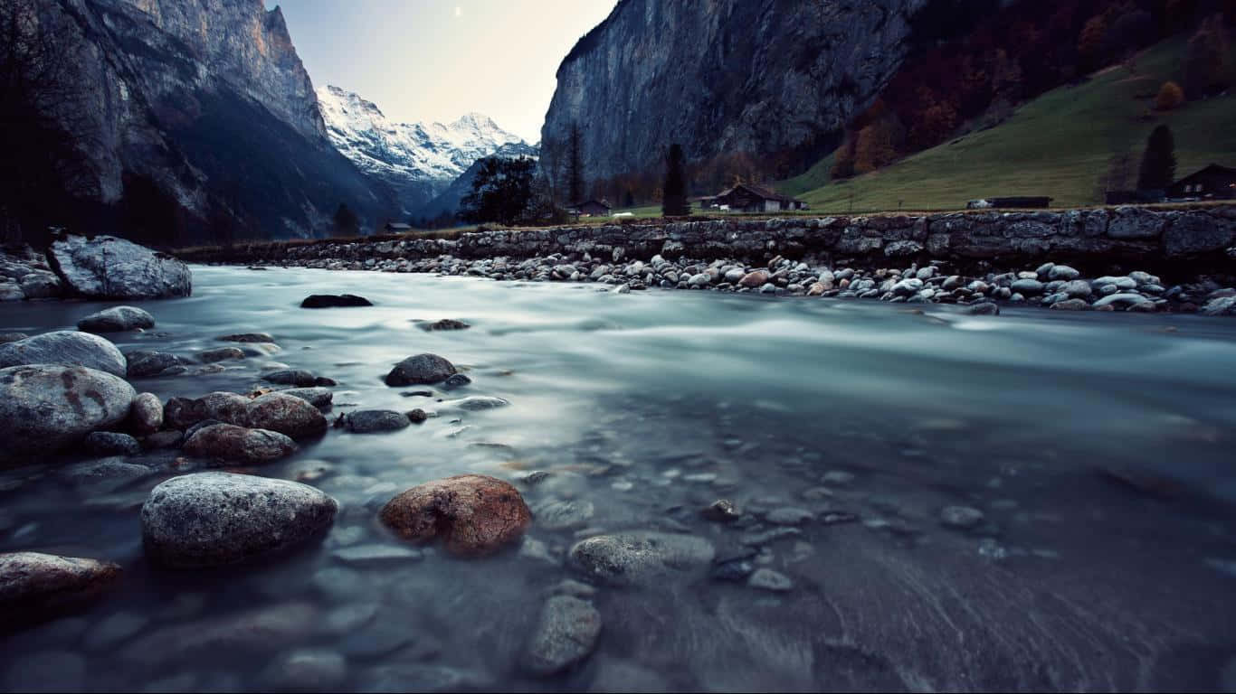 溪流山景 1366x768 顯示器背景 壁紙 — 一條寧靜的山間溪流流過光滑的石頭，遠處有積雪的山峰和一座石屋。