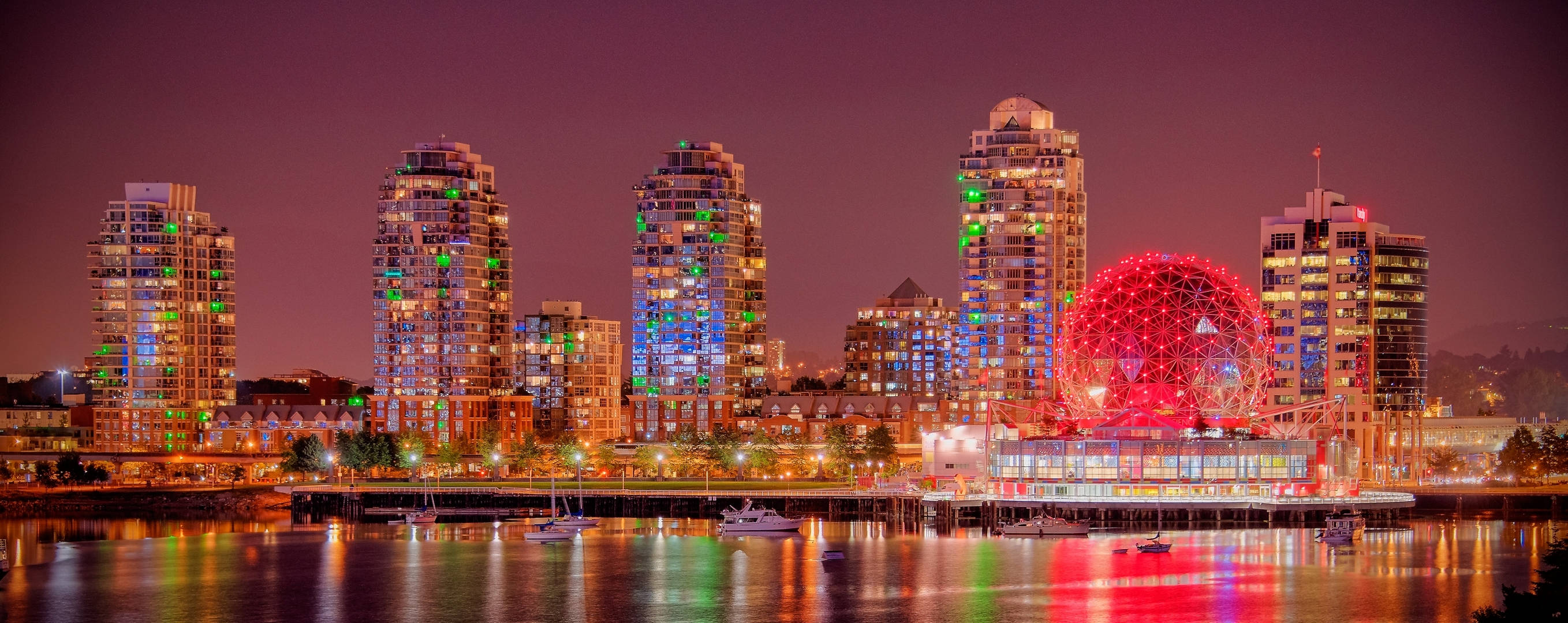 溫哥華城市燈光 壁紙 — 充滿活力的城市天際線夜景，色彩繽紛的摩天大樓和發光的紅色球形結構倒映在平靜的水面上。