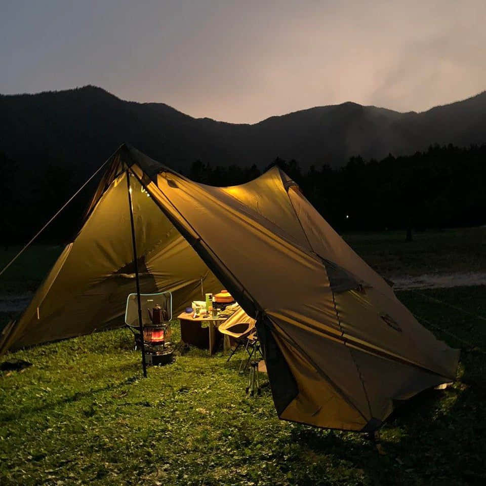 Minimalist Camping 桌布 壁紙 — 溫暖照亮的帳篷在黑暗的山巒映襯下閃耀，營火和桌子邀請您在暮色中享受舒適的孤獨。
