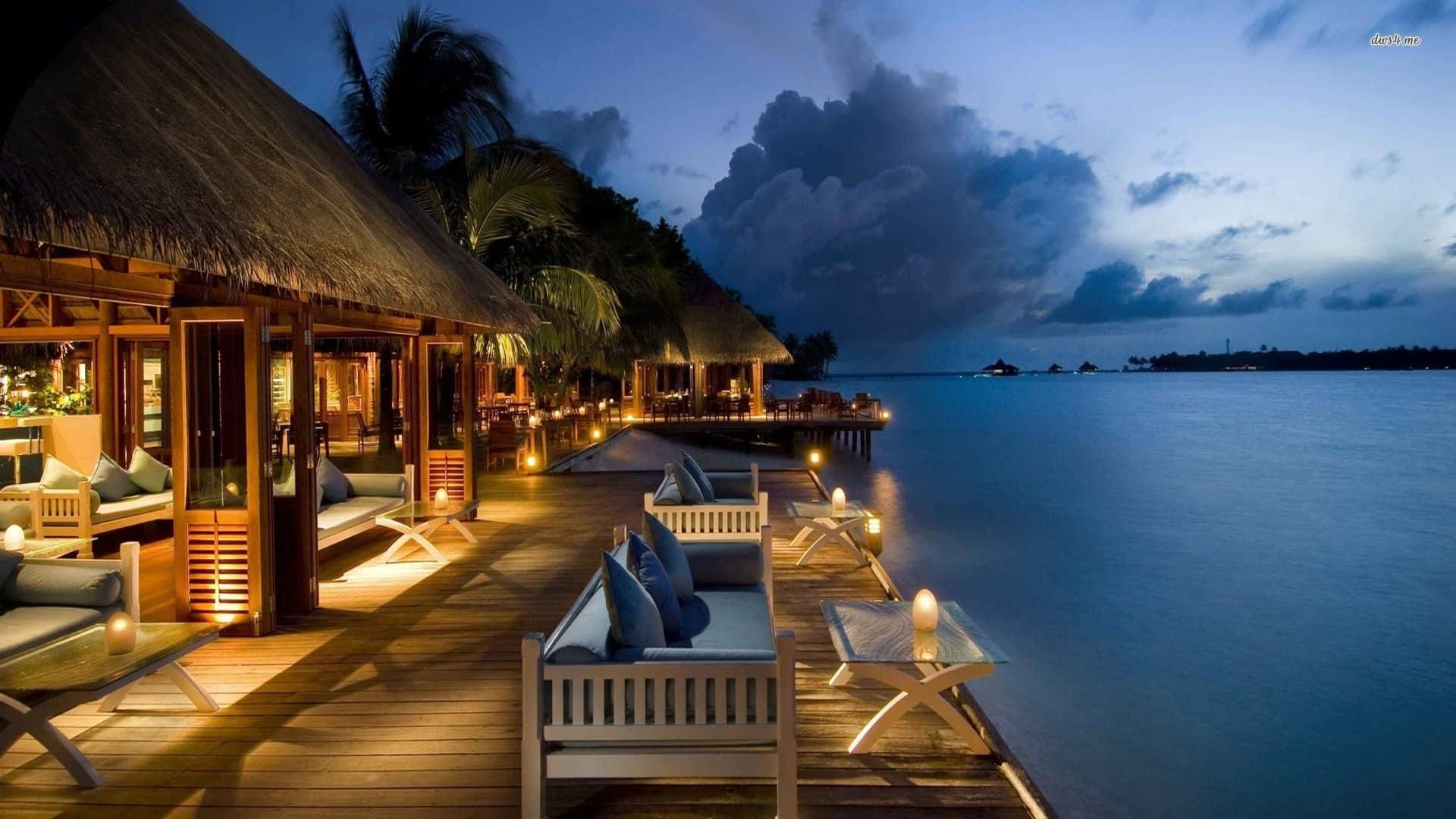 Maldives Beach 桌布 壁紙 — 溫暖照明的木質甲板，茅草屋頂小屋和躺椅延伸至暮色天空下平靜的海洋之上。