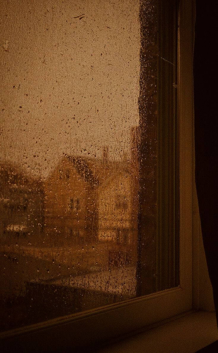 Raindrops 桌布 壁紙 — 溫暖的室內光線穿過佈滿雨點的窗戶，映出外面金色暮色中模糊的磚砌建築。