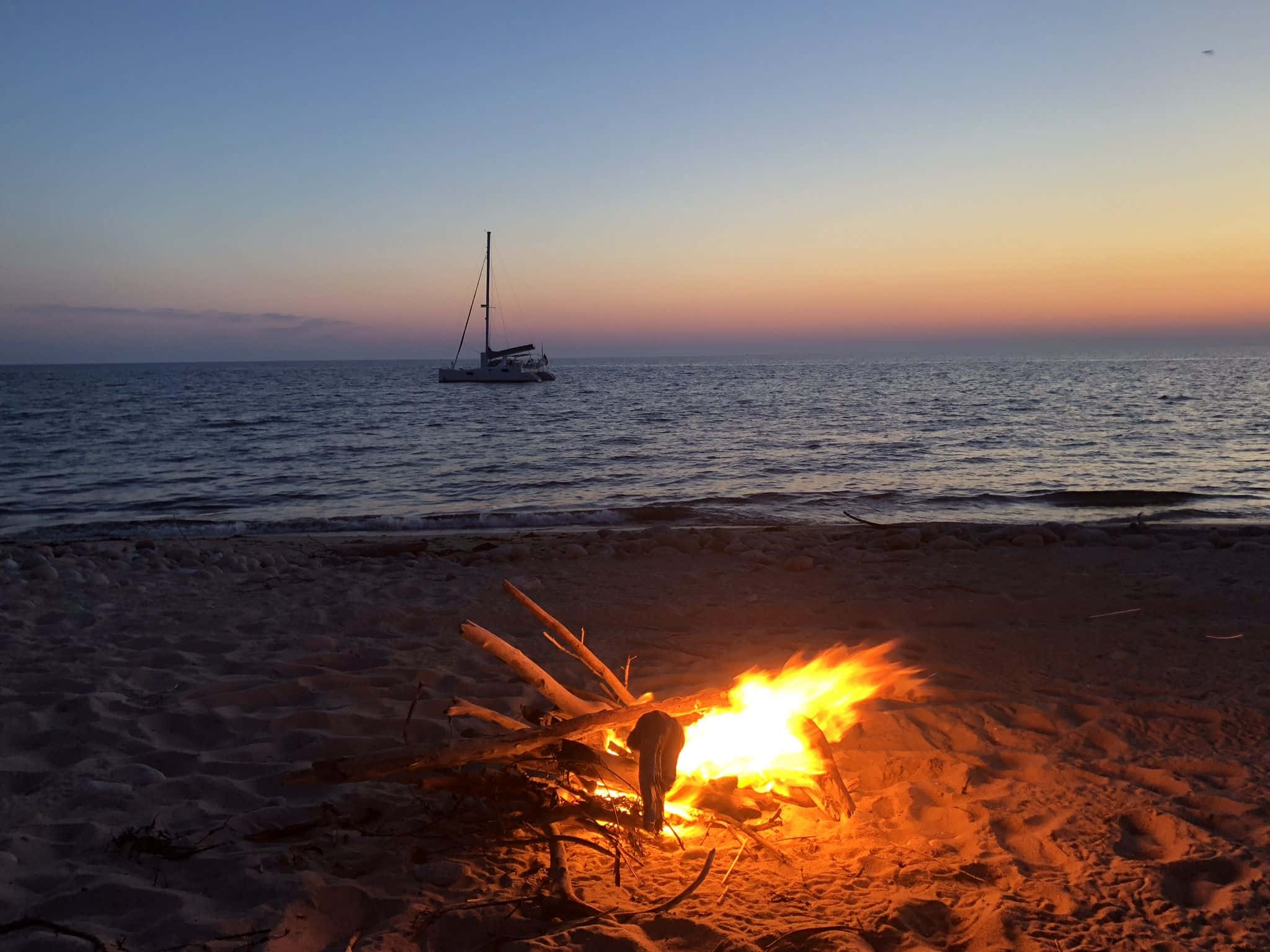Beach Bonfire 桌布 壁紙 — 溫暖的營火在沙灘上閃耀，日落時分，一艘帆船在平靜的海平線上形成剪影。