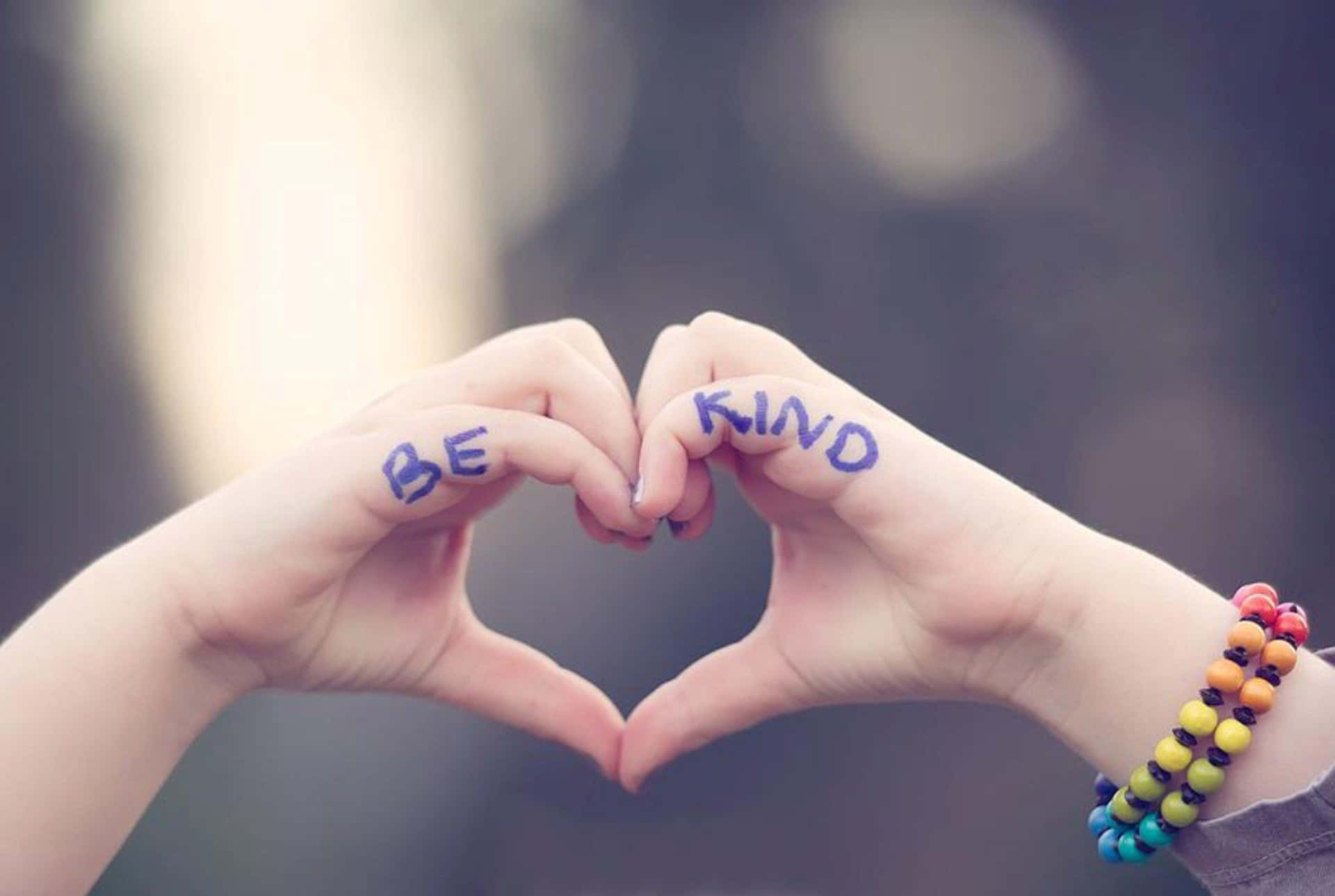 兩隻手形成一個心形，手掌上寫著「BE KIND」，一隻手腕上戴著彩色手鐲。