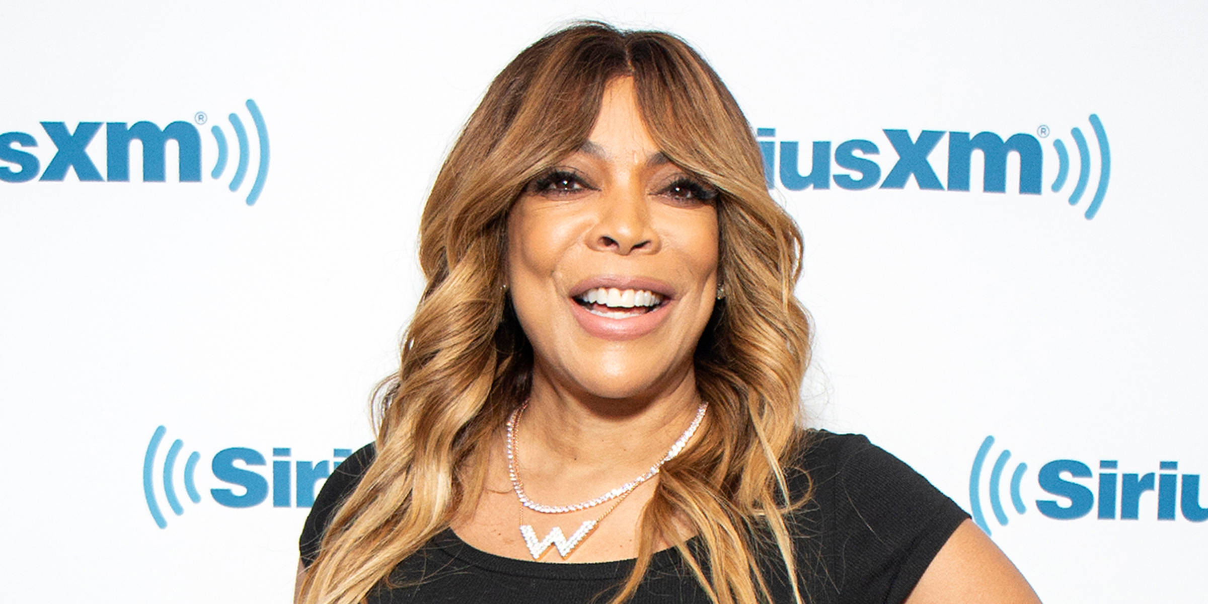 溫迪·威廉姆斯佩戴W字項鍊 壁紙 — Wendy Williams 穿著黑色上衣和閃亮的W項鍊，在SiriusXM品牌背景前對著鏡頭燦爛微笑。