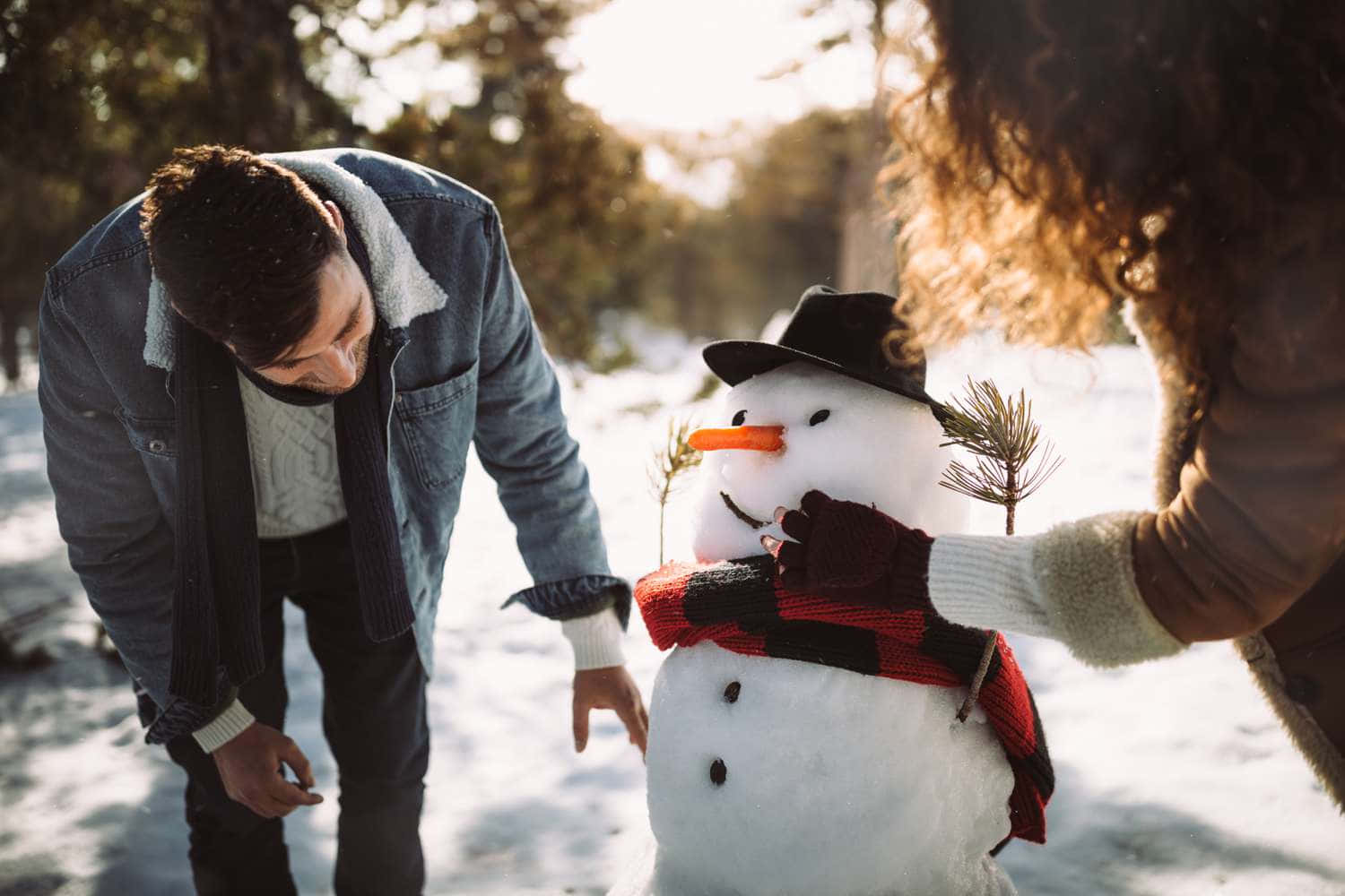 溫馨冬日風情隨拍：情侶與雪人 壁紙 — 一對情侶穿著保暖的冬季服裝，在陽光明媚的雪地森林中，輕輕地用圍巾和帽子裝飾雪人。