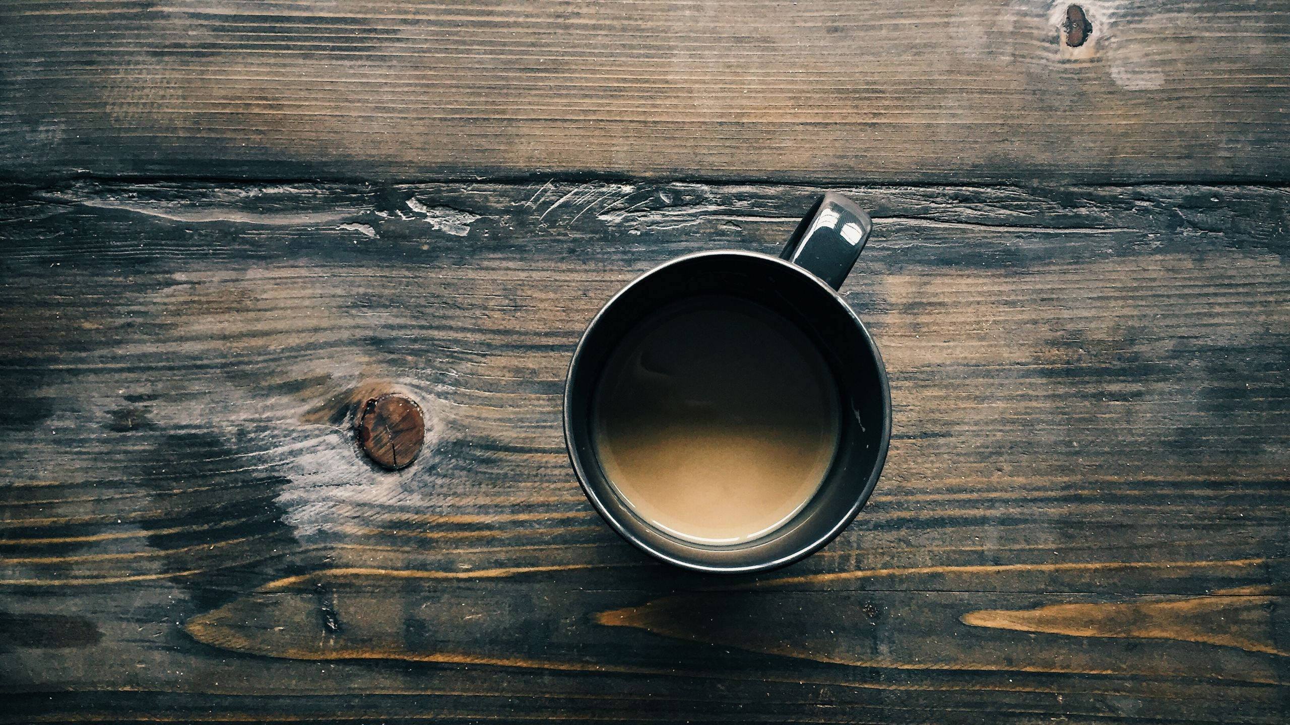 溫馨咖啡杯木質桌面 壁紙 — 一個裝滿咖啡的黑色馬克杯放在一張風化的深色木桌上，木紋和節疤清晰可見。