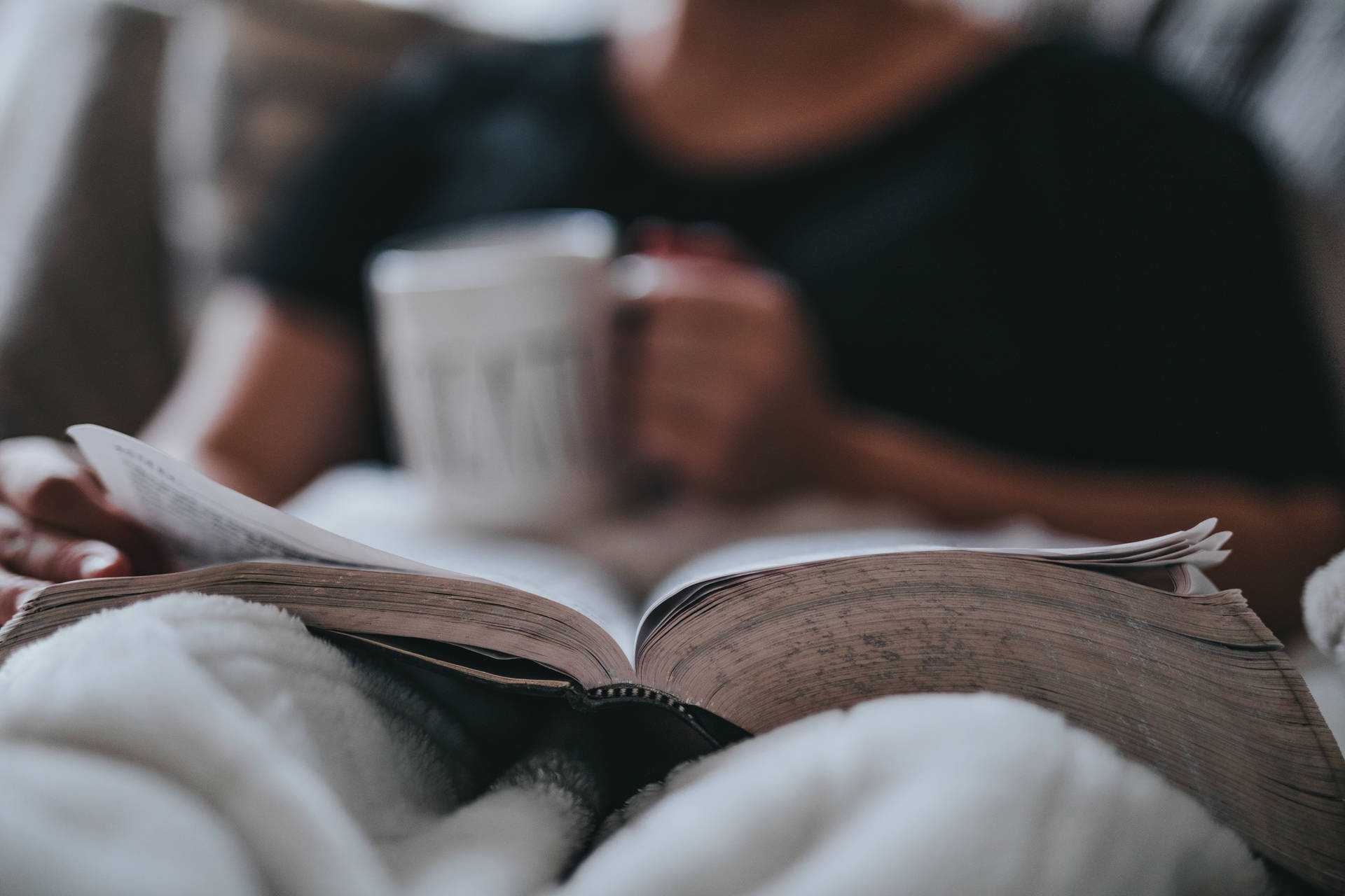 一個人在柔軟毯子下拿著杯子閱讀一本書。